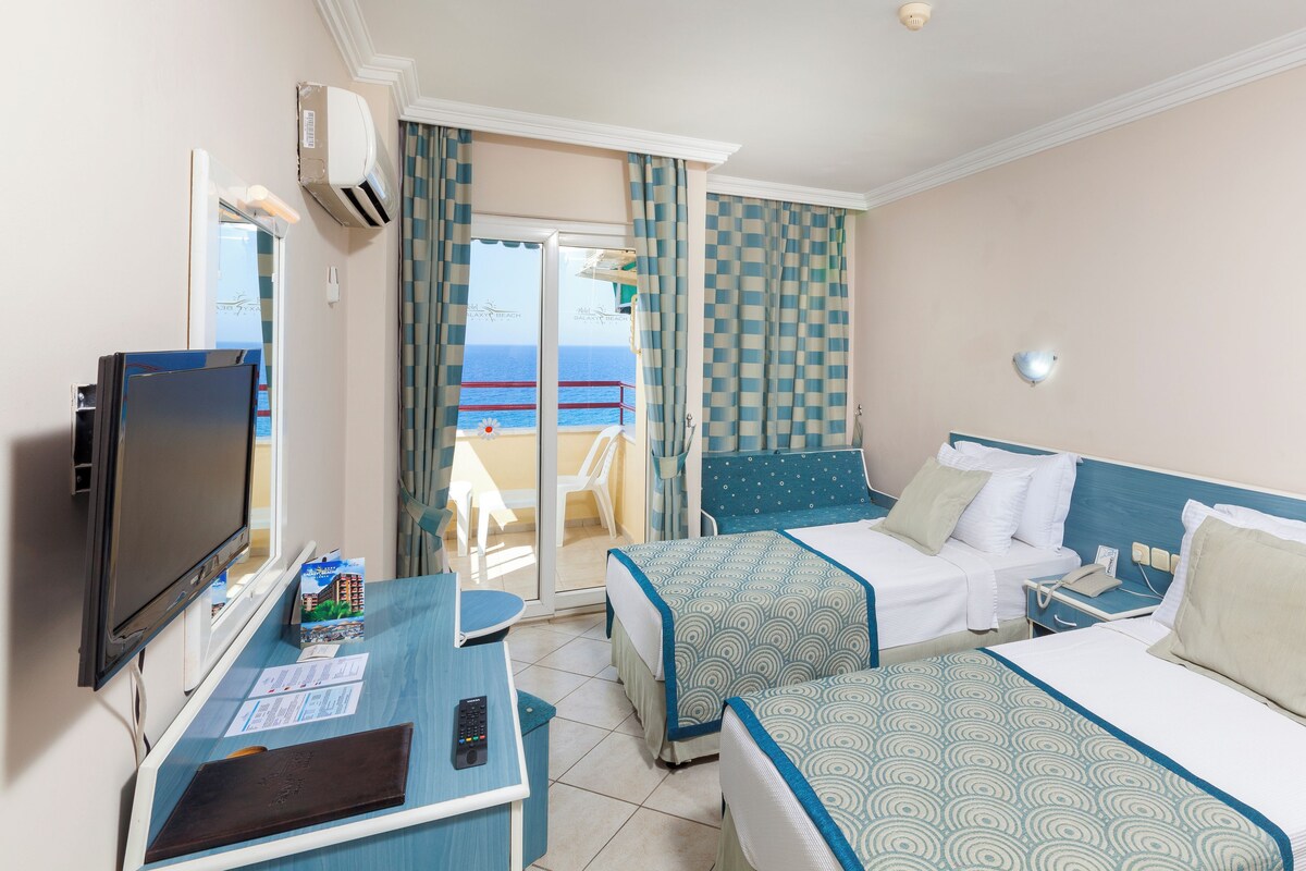 A hotel room with two beds, a TV, a desk with a remote control, and a view of the sea.