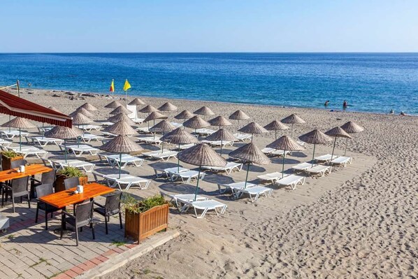 A beachside area with sun loungers and umbrellas, a wooden table with chairs, and a view of the sea.