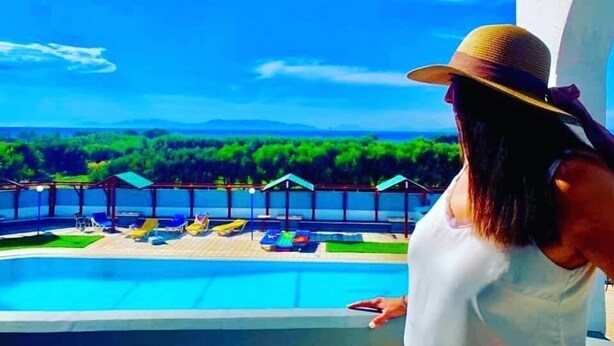 Une personne se tient au bord d’une piscine, entourée de chaises longues et de parasols, avec une végétation luxuriante et un ciel dégagé en arrière-plan.