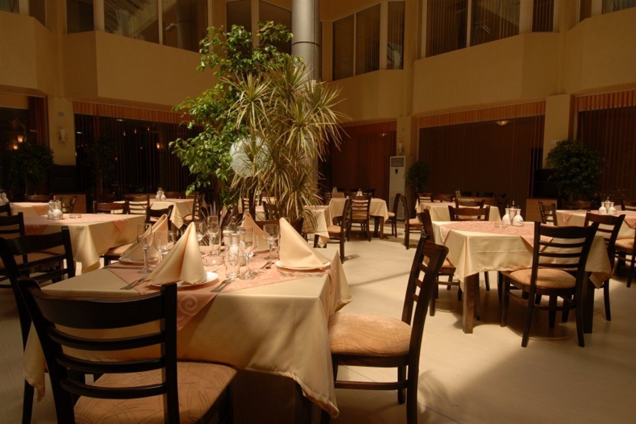 A spacious indoor dining area with multiple tables set for guests, featuring a central decorative plant and a high ceiling with large windows.