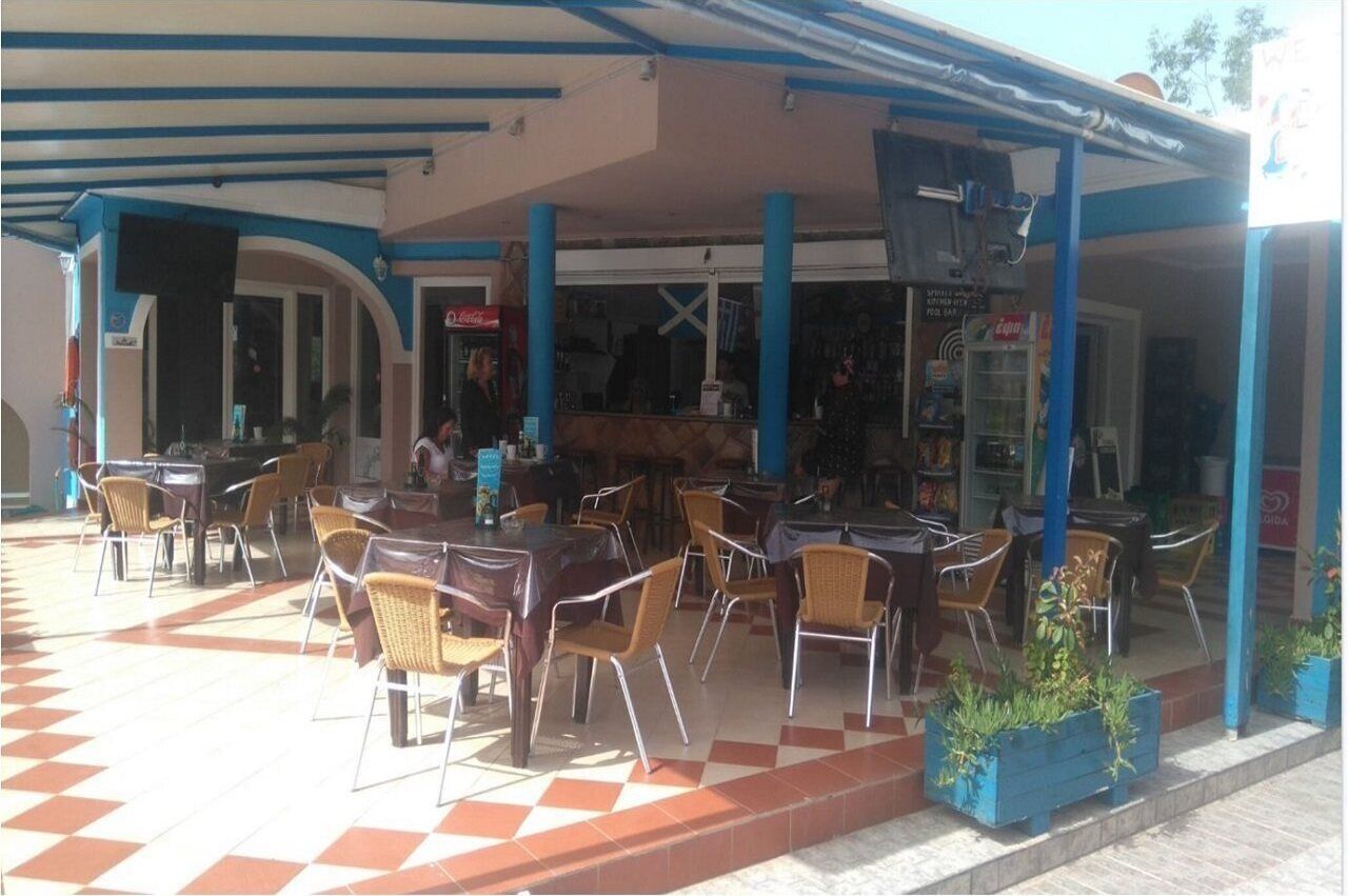 An outdoor cafe with tables and chairs, a covered seating area, and a storefront with visible signage.