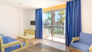 Una habitación con sofá, una mesita, televisor y balcón con cortinas azules.