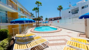 Un área de piscina con tumbonas y sombrillas, un edificio con balcones y palmeras.
