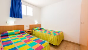 Una habitación con dos camas, cada una con un edredón de colores, una ventana con cortinas azules y una mesita de noche con lámpara.