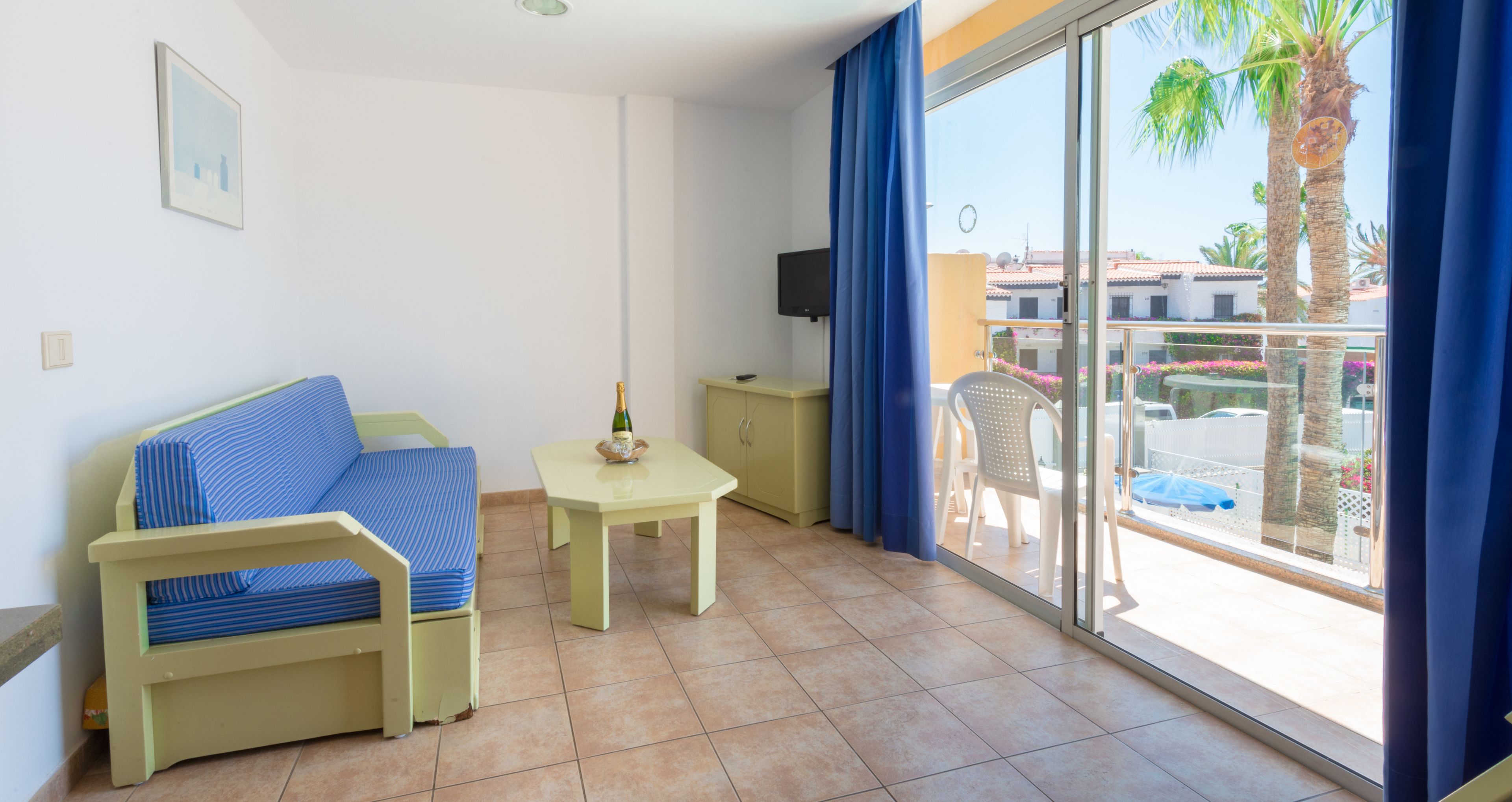Une pièce avec un canapé, une petite table sur laquelle se trouve une bouteille, un téléviseur fixé au mur et un balcon avec des chaises et un palmier.