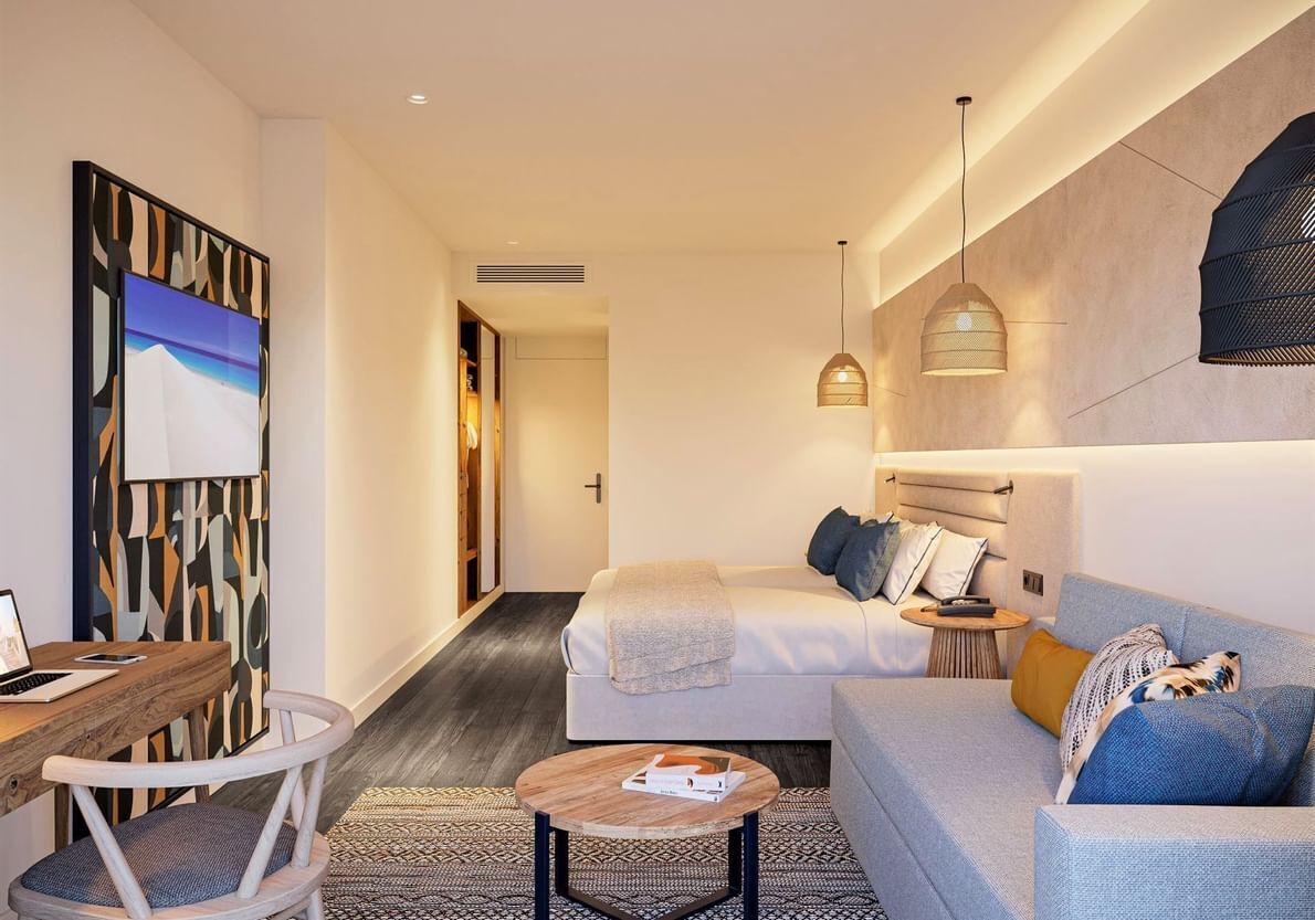 A modern hotel room with a bed, a sofa, a desk, and a chair. There is a painting on the wall and a hanging light fixture.