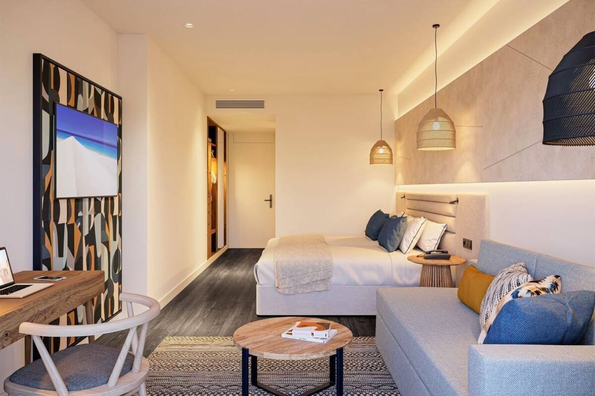 A modern hotel room with a bed, a sofa, a desk, and a chair. There is a painting on the wall and a hanging light fixture.