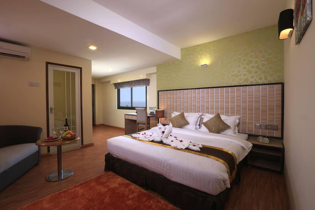 Ein Hotelzimmer mit einem großen Bett, einem Schreibtisch mit Stuhl, einem kleinen Tisch mit Obstschale und Weingläsern sowie einem Sofa.