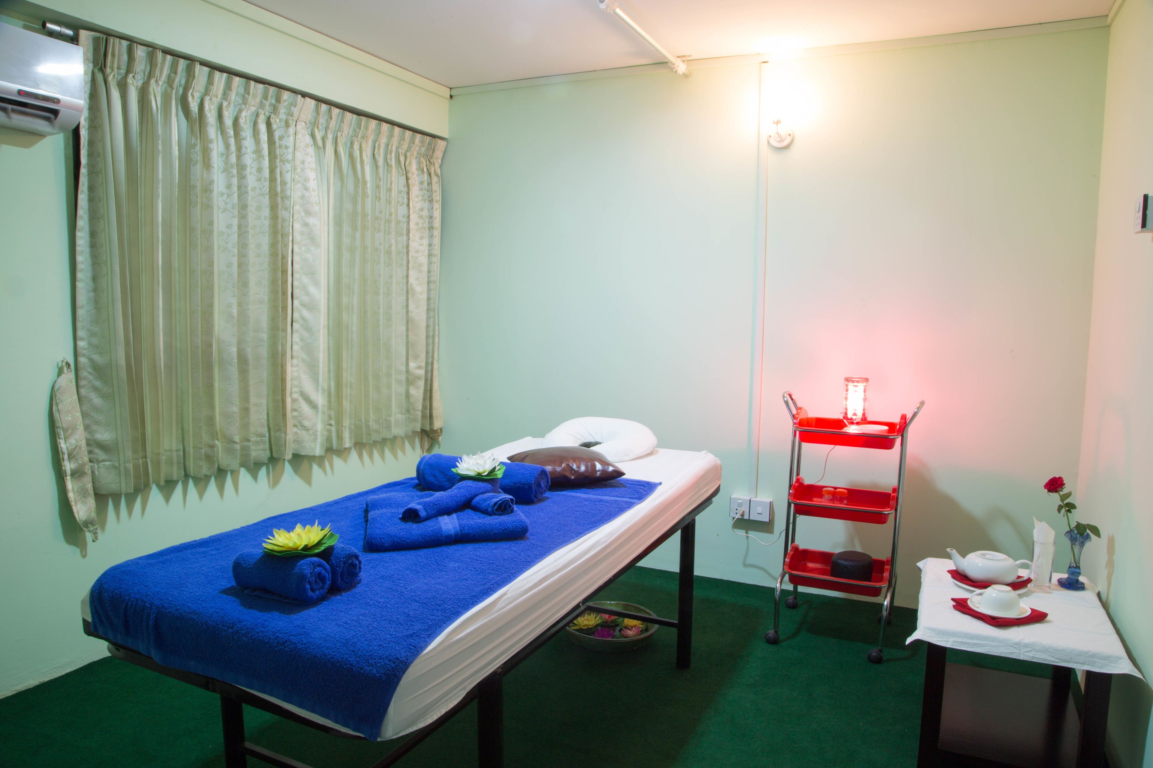 A room with a bed, a small table with a vase and a rose, a red cart with a light, and a curtain.