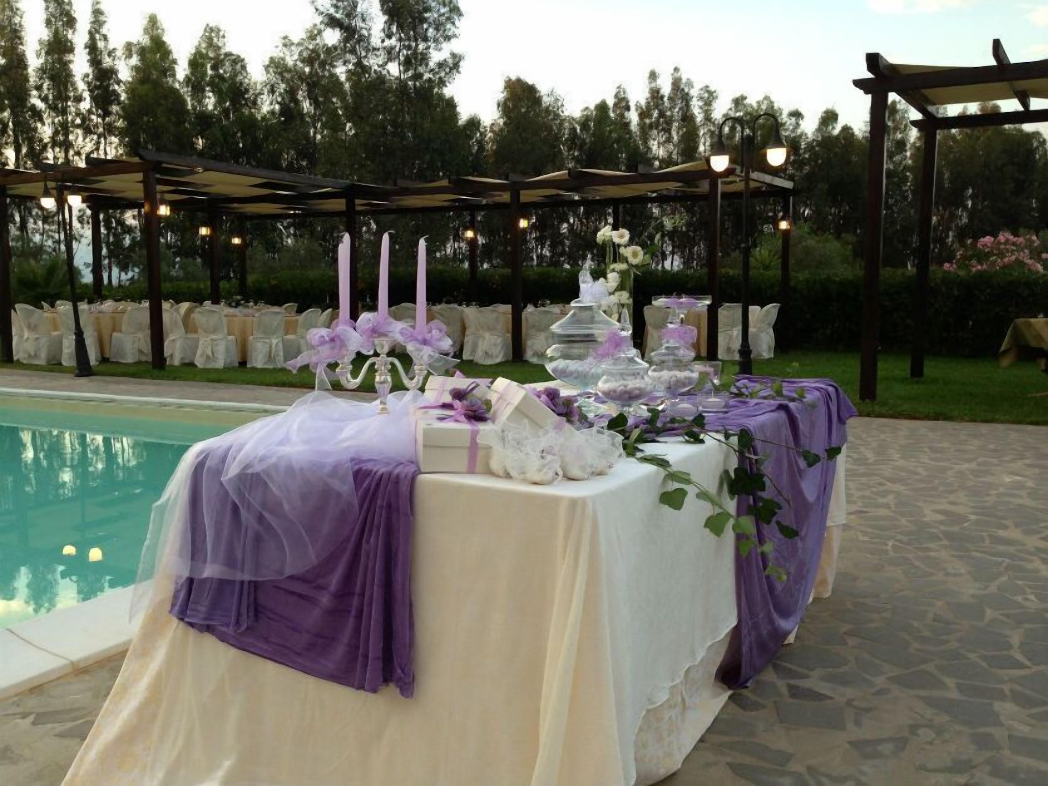 An outdoor wedding setup with a table covered in white and purple, surrounded by chairs and a pergola.