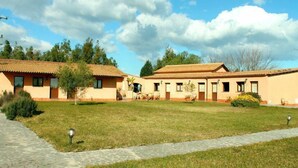 A courtyard with a paved walkway, a grassy area, and a building with a tiled roof and beige walls. - Le Terre di San Domenico (Augusta)