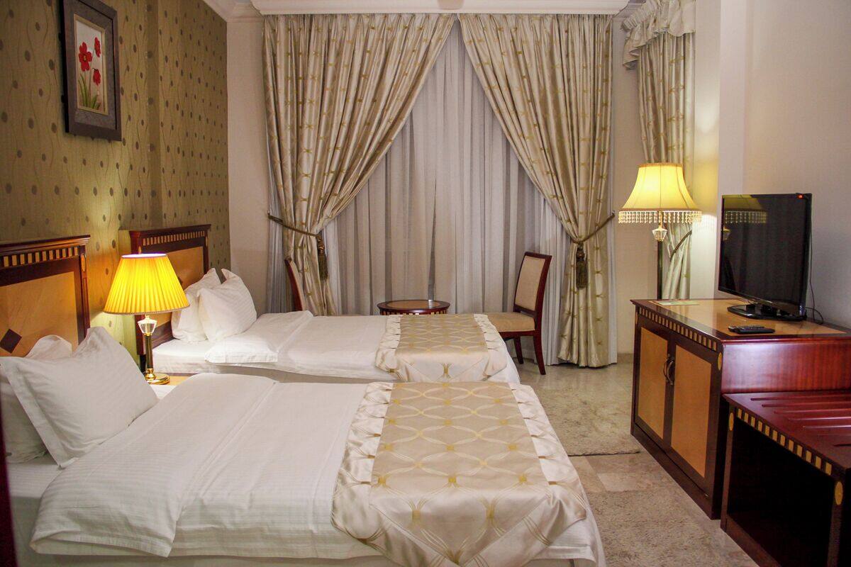 A hotel room with two beds, a desk, a chair, a TV, and a lamp.