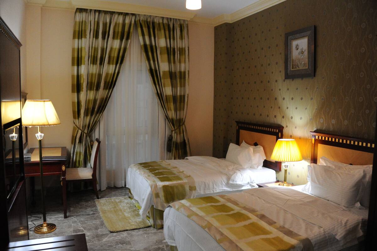 A hotel room with two beds, a desk with a lamp, a chair, and a window with curtains.