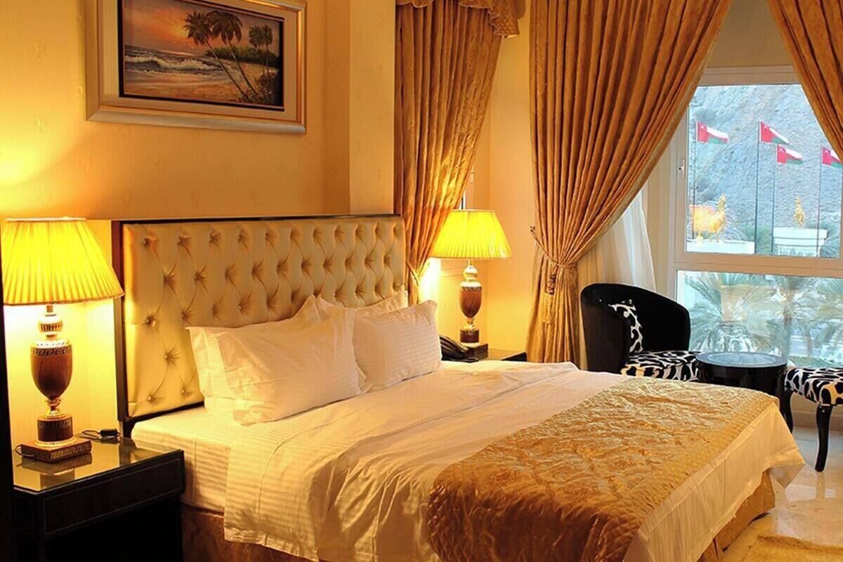 A hotel room with a large bed, two bedside lamps, a framed picture on the wall, and a view of palm trees through the window.