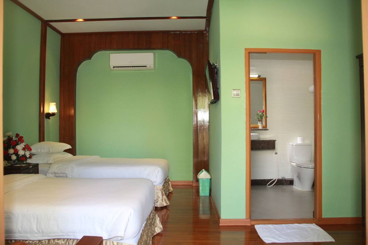 Une chambre d’hôtel avec deux lits, un plancher en bois, un appareil de climatisation, une salle de bain avec des toilettes et un lavabo, et une petite poubelle.