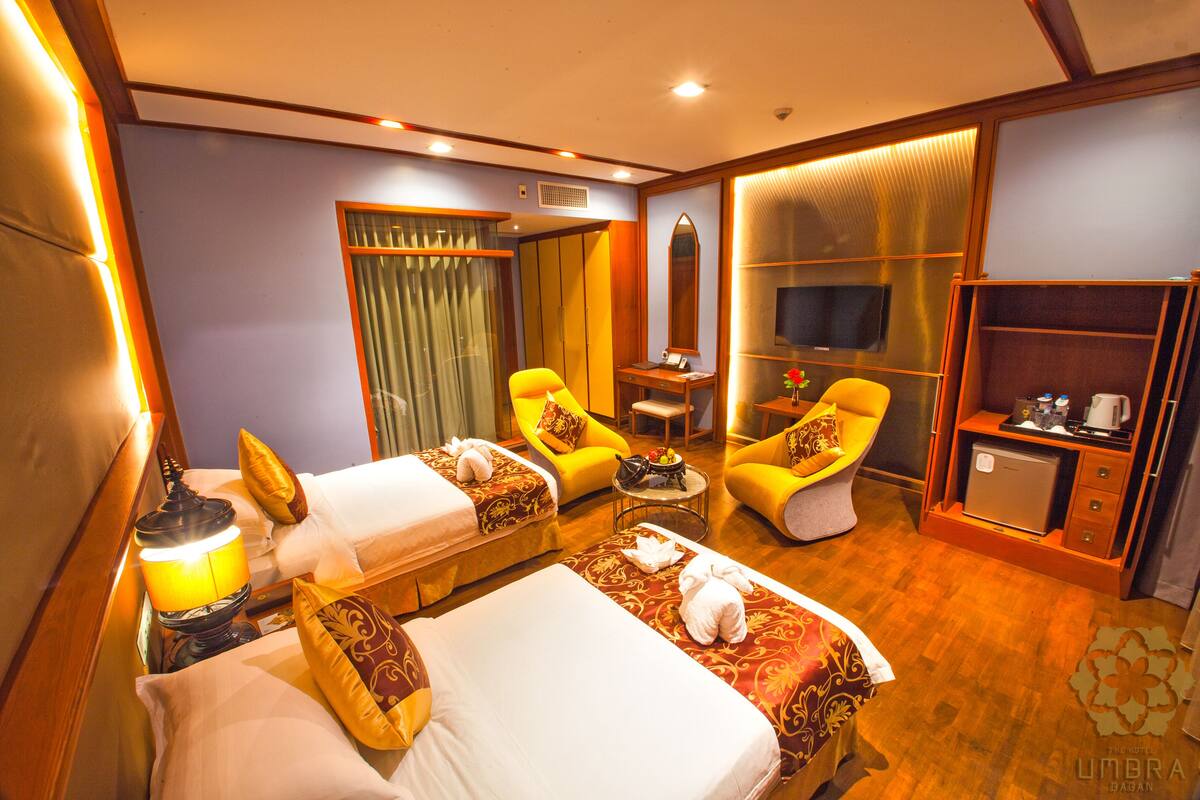 Une chambre d’hôtel avec deux lits, un coin salon comprenant un canapé et des fauteuils, une petite table avec une lampe, une télévision et un mini-bar.
