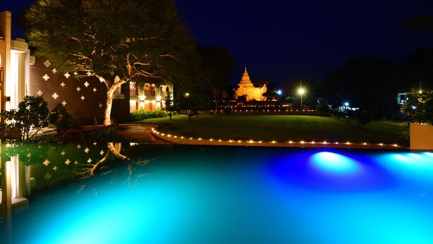 Vista nocturna de un hotel con piscina, un árbol grande y un templo al fondo.
