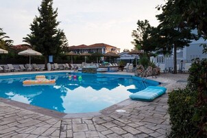 Una piscina con sillones de descanso y sombrillas, rodeada de árboles y un edificio al fondo.