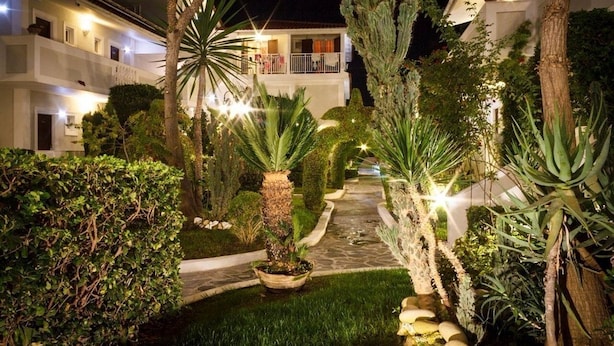 Caminho iluminado em um jardim que leva a um edifício com várias varandas, rodeado por plantas e árvores diversas.
