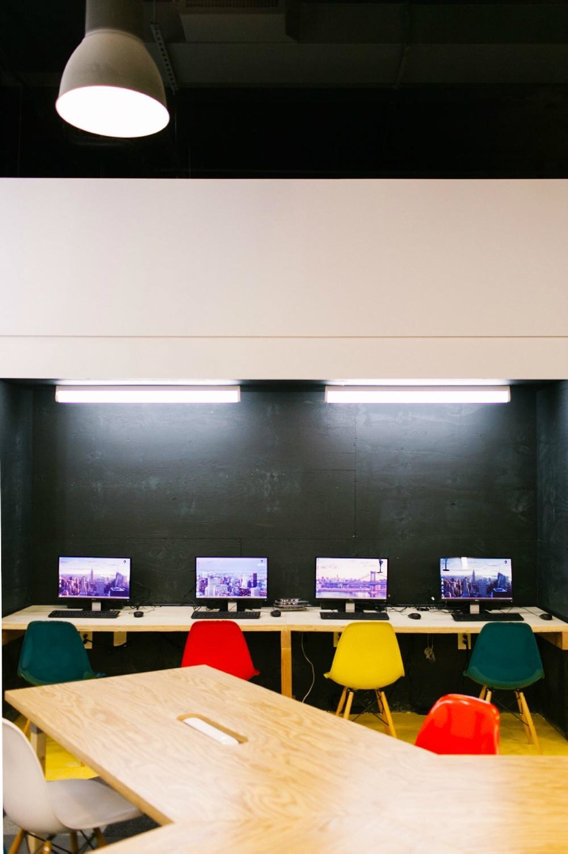 Un salón de conferencias con una larga mesa, sillas coloridas y cuatro monitores de computadora en una pared oscura.