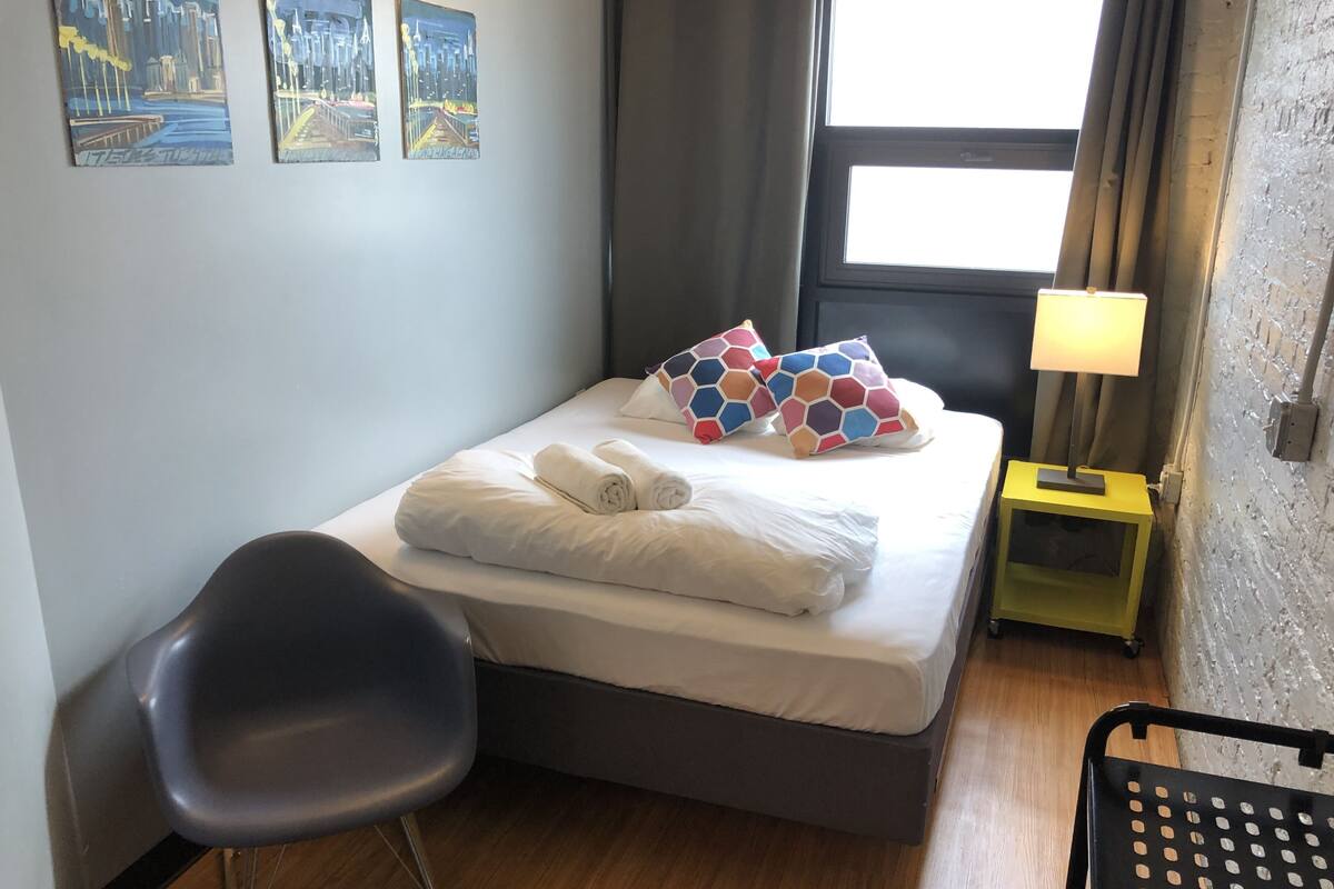 A neatly made bed with two pillows, a bedside table with a lamp, a chair, and artwork on the wall.