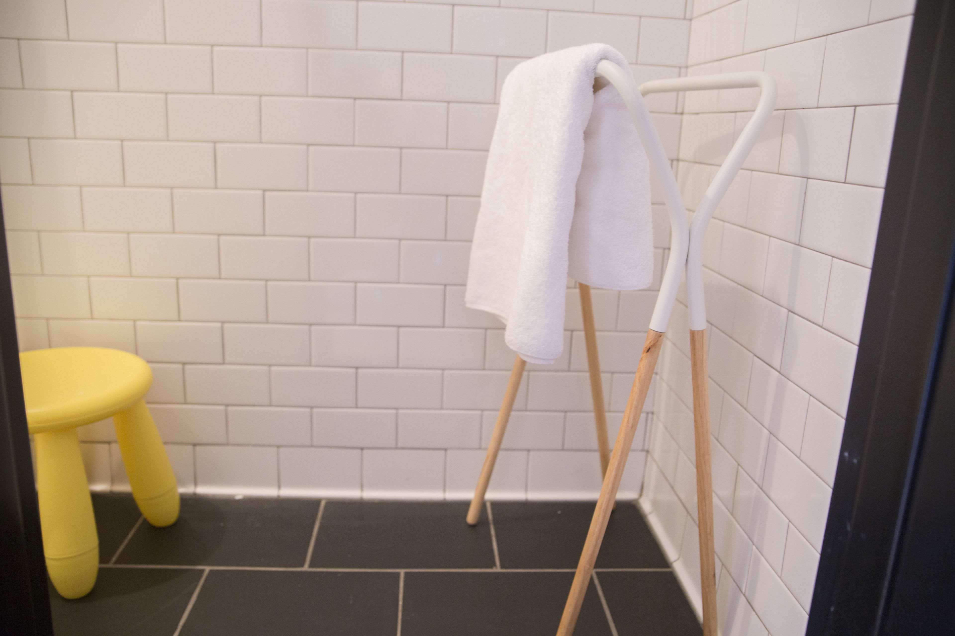 A bathroom with white tiled walls, a black floor, a white towel on a wooden towel rack, and a yellow stool.