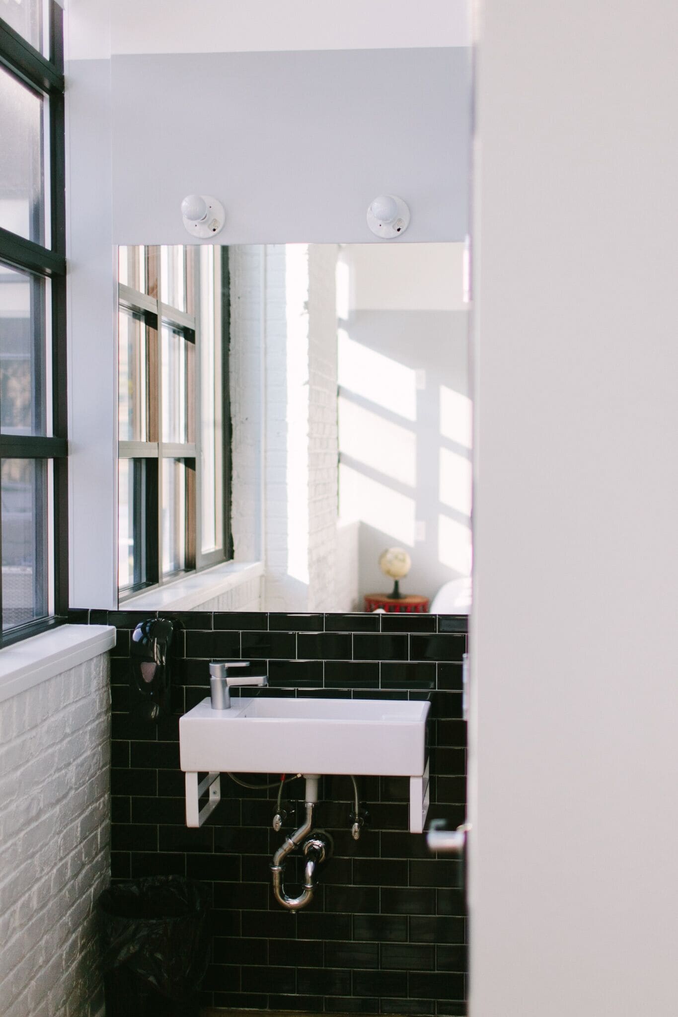 A bathroom with a white sink, black tiled walls, a window with blinds, and a globe on a small table.