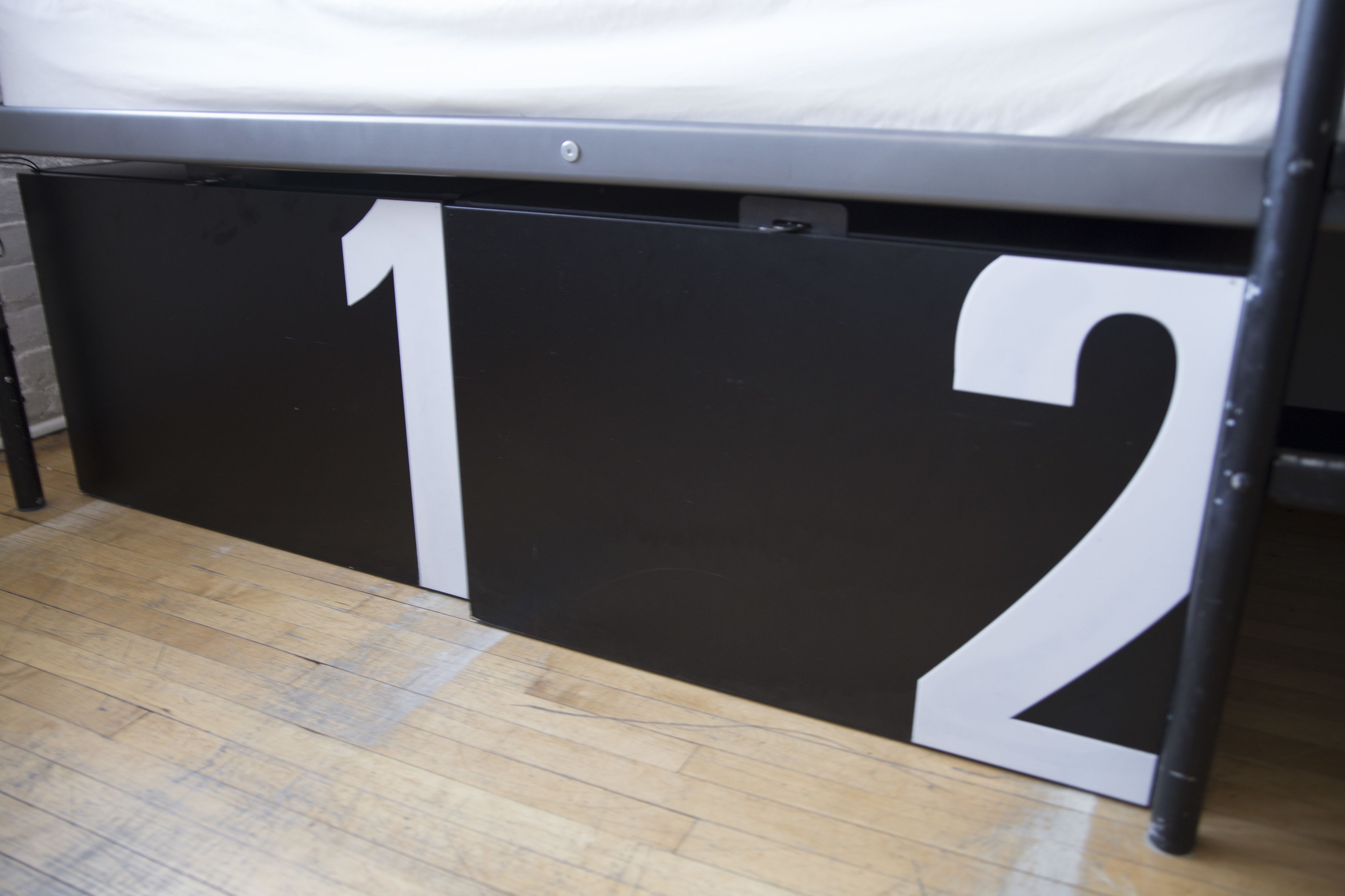 Een bed met een zwart metalen frame en een witte matras. Het bed staat op een houten vloer, waarop het cijfer '12' zichtbaar is.