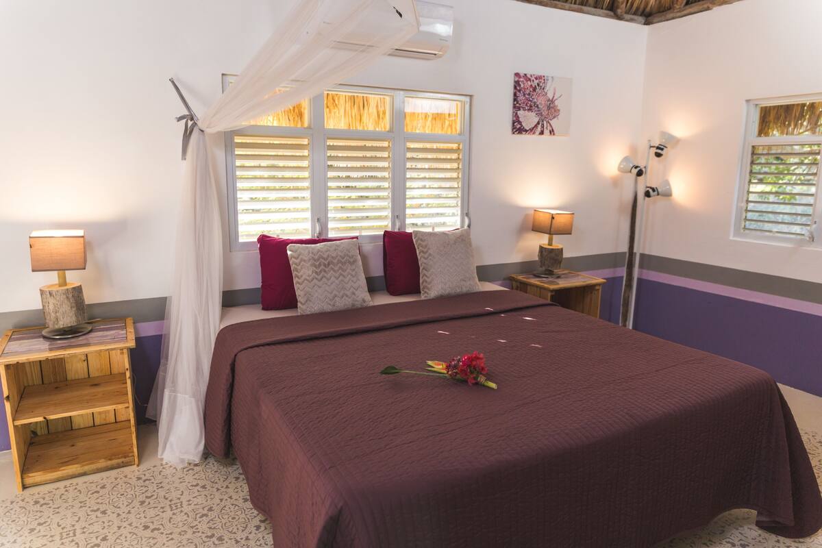 A bedroom with a large bed, a bedside table with a lamp, a window with shutters, and a wall-mounted artwork.