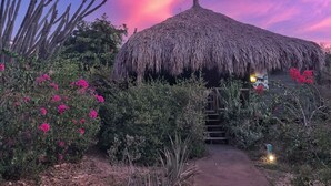Una cabaña con techo de paja, con un sendero de tierra que la lleva a ella, rodeada de abundante vegetación y flores rosadas.