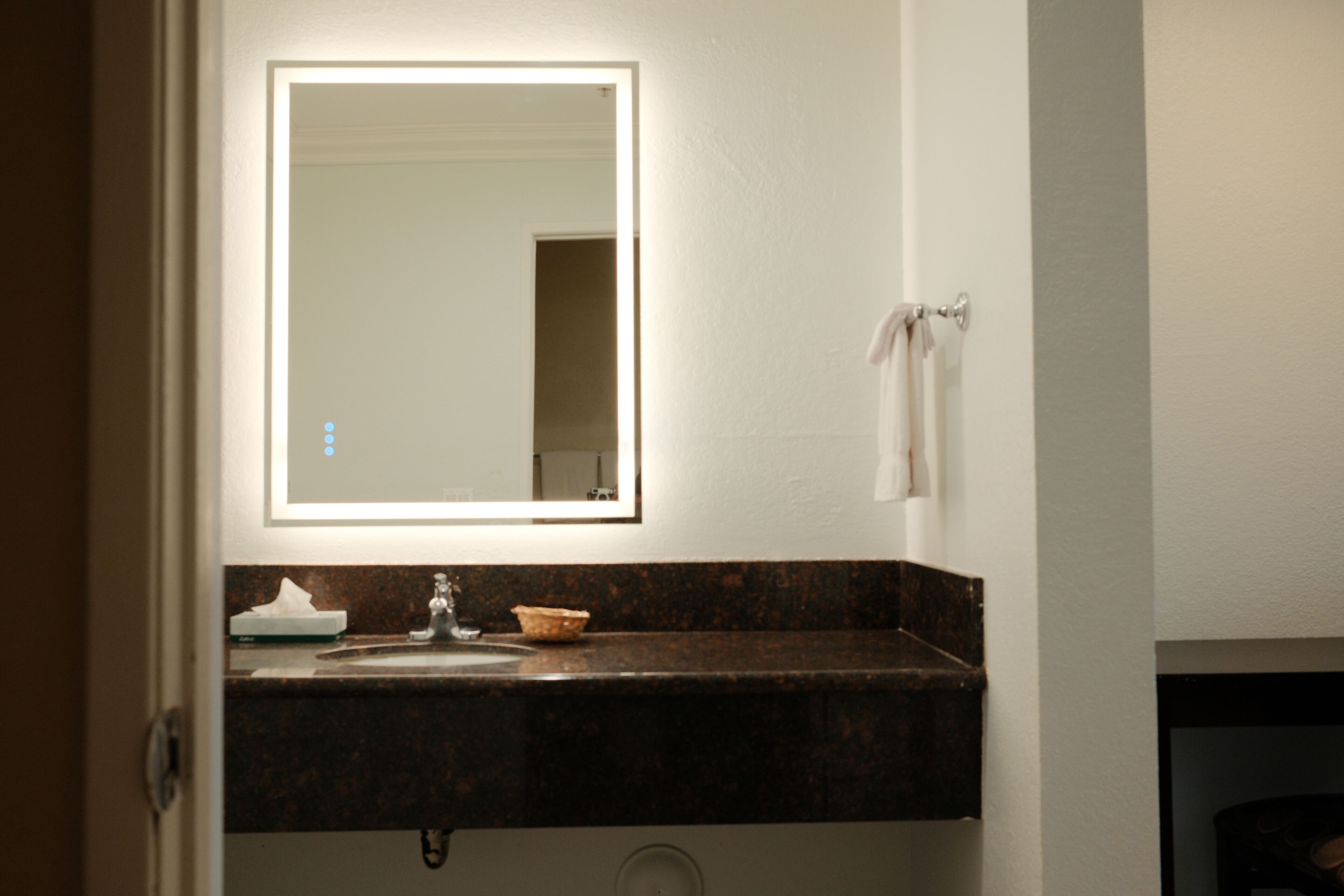 A bathroom with a marble countertop, a sink, a mirror with a light, and a towel hanging on a hook.