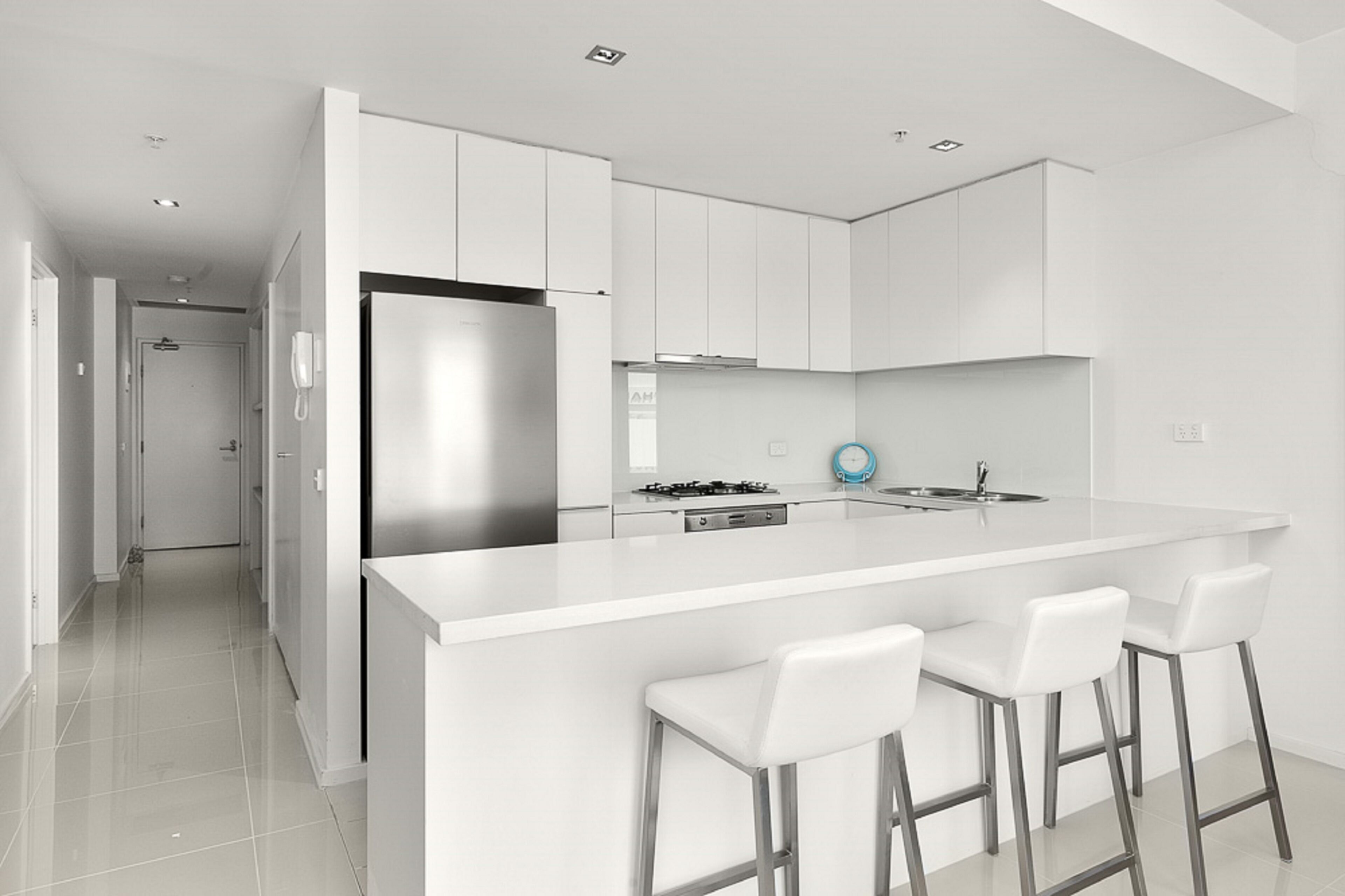 A modern kitchen with white cabinetry, a stainless steel refrigerator, a gas stove, and a bar area with white stools.