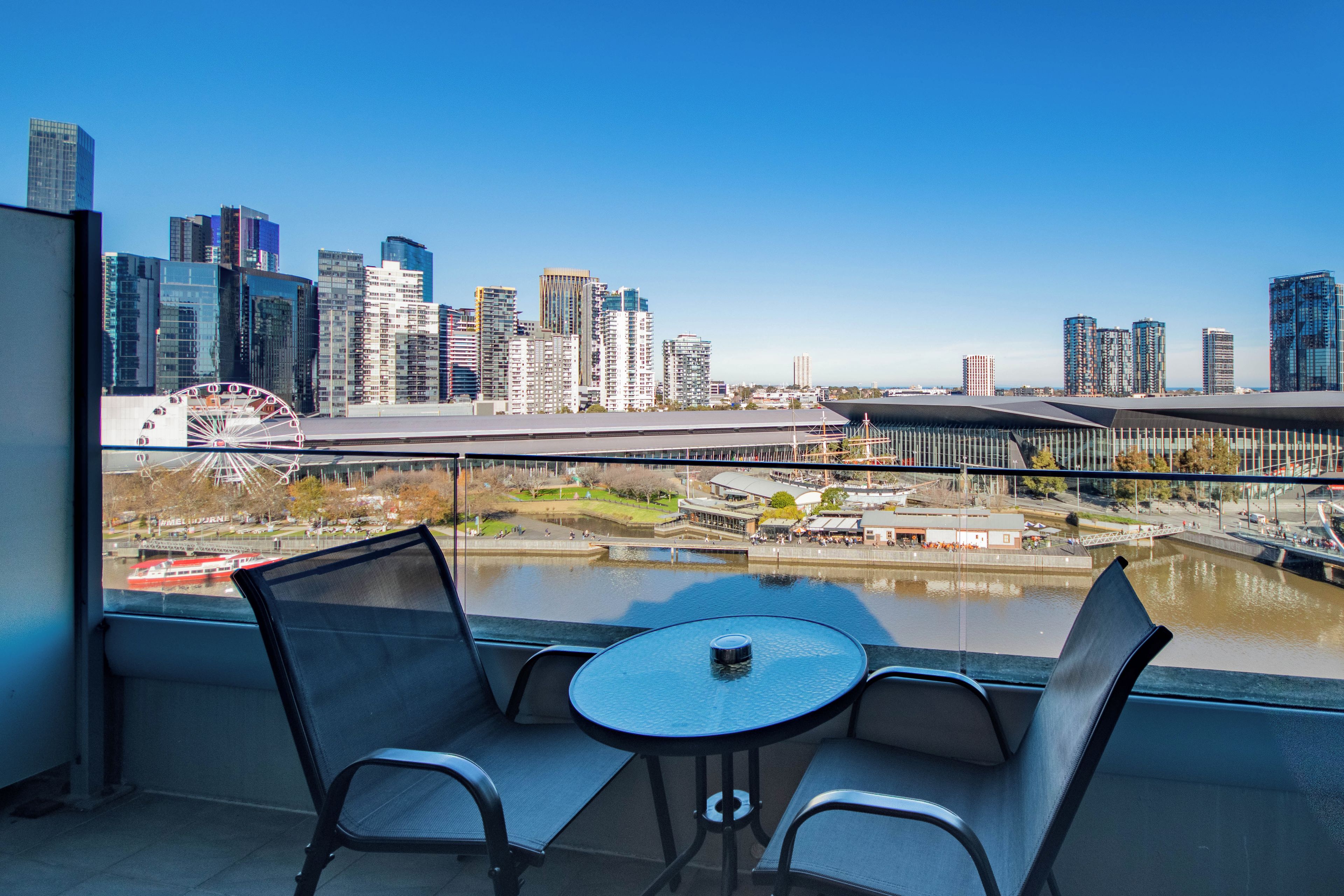Un balcon avec une table et des chaises donnant sur un paysage urbain, avec des bâtiments modernes et une grande roue.