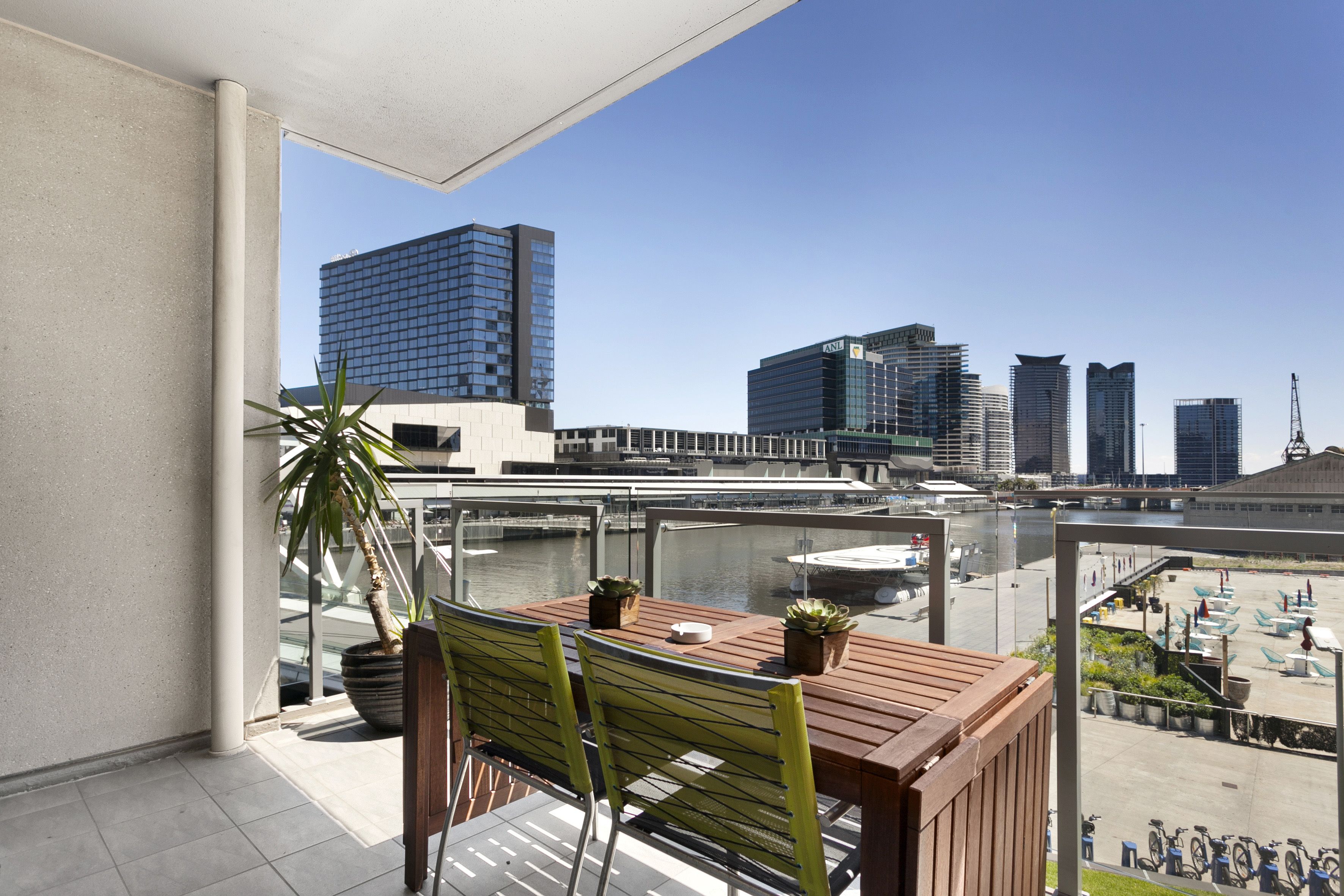 A balcony with a wooden table and chairs, potted plants, and a view of a cityscape with modern buildings.