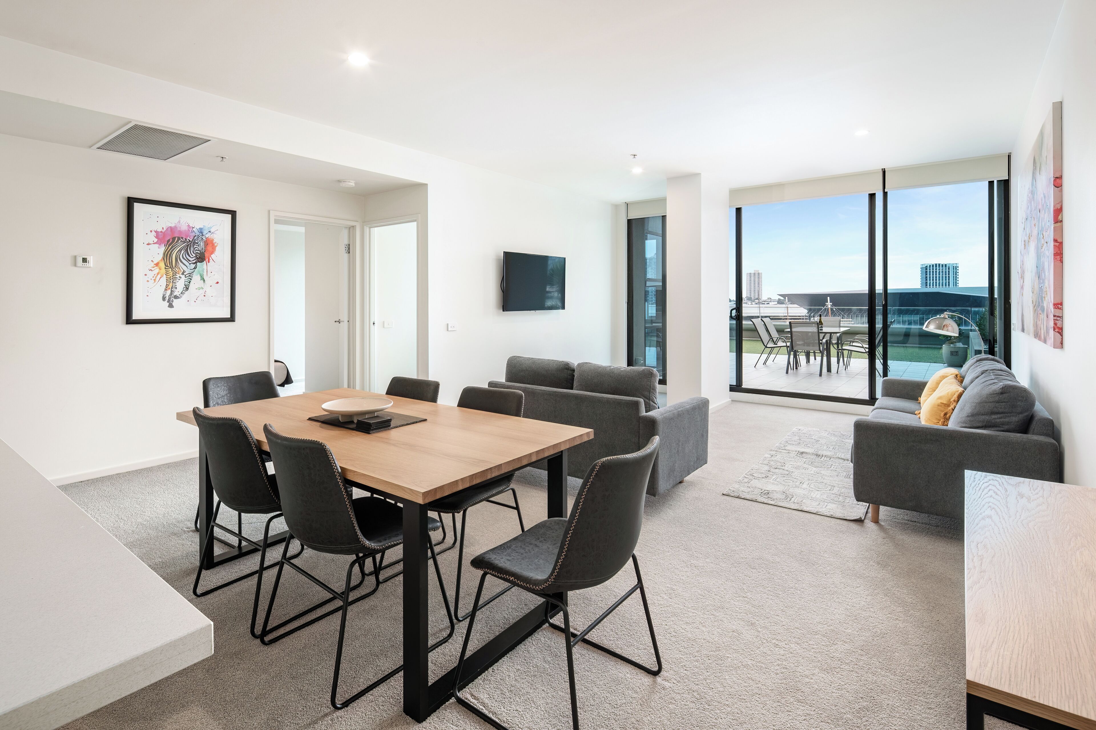 A modern living room with a wooden dining table, grey sofas, and a large window offering a city view.