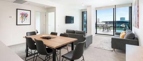 A modern living room with a wooden dining table, grey sofas, and a large window offering a city view.