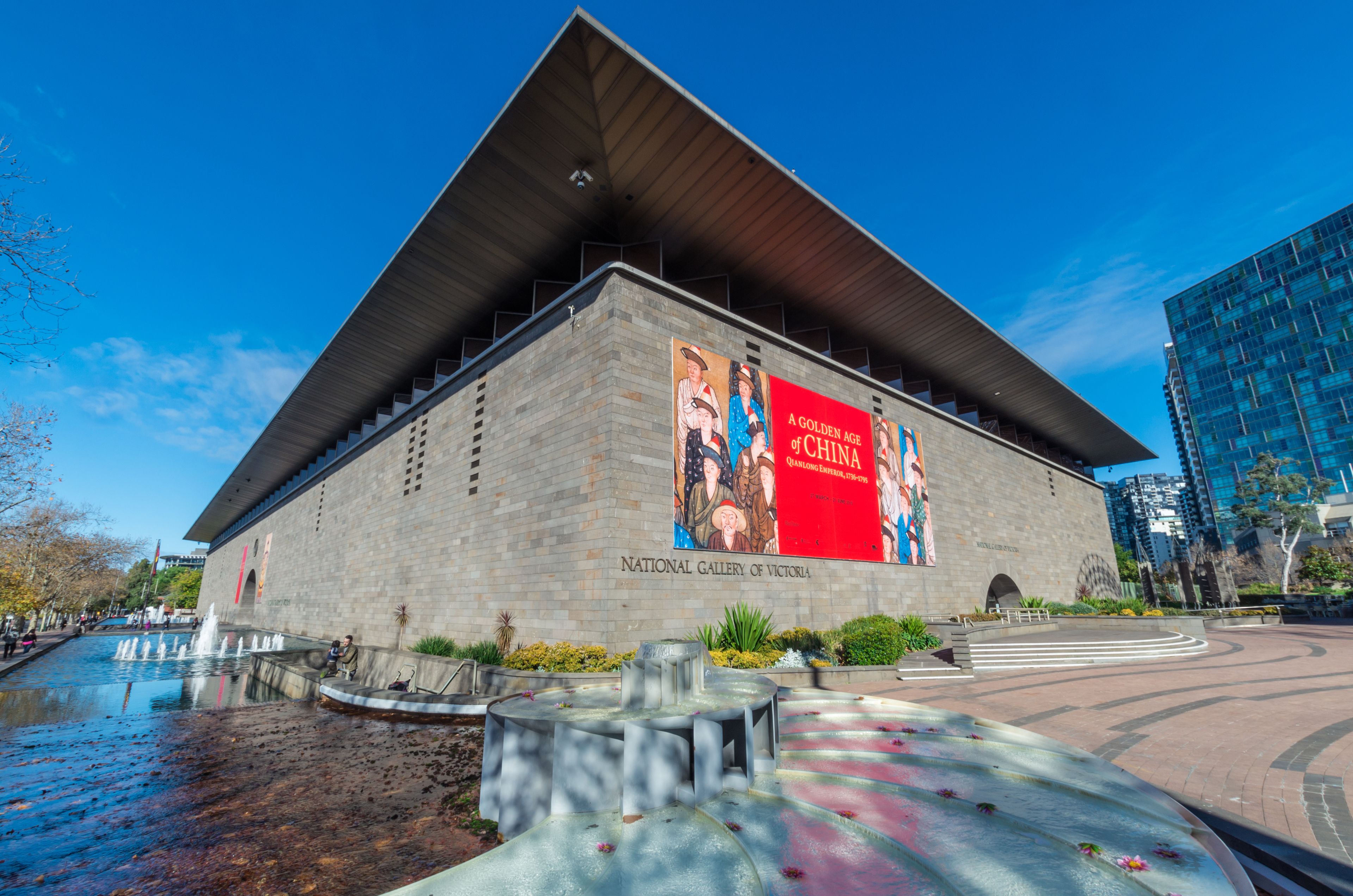 La Galería Nacional de Victoria, con un diseño arquitectónico moderno, presenta un gran cartel rojo y un elemento decorativo de agua al frente.