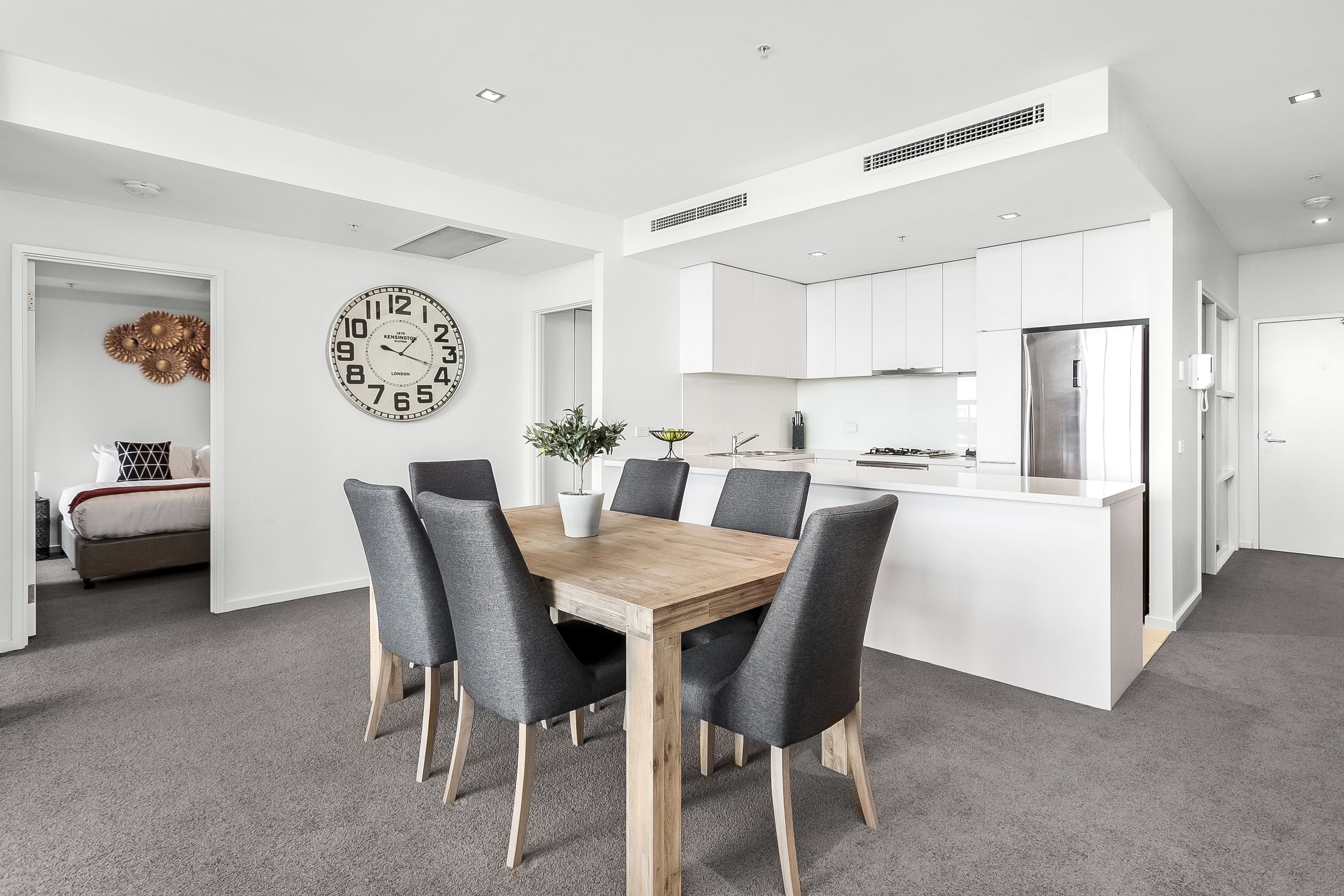 A modern kitchen with white cabinetry, a stainless steel refrigerator, and a dining area with a wooden table and grey upholstered chairs.