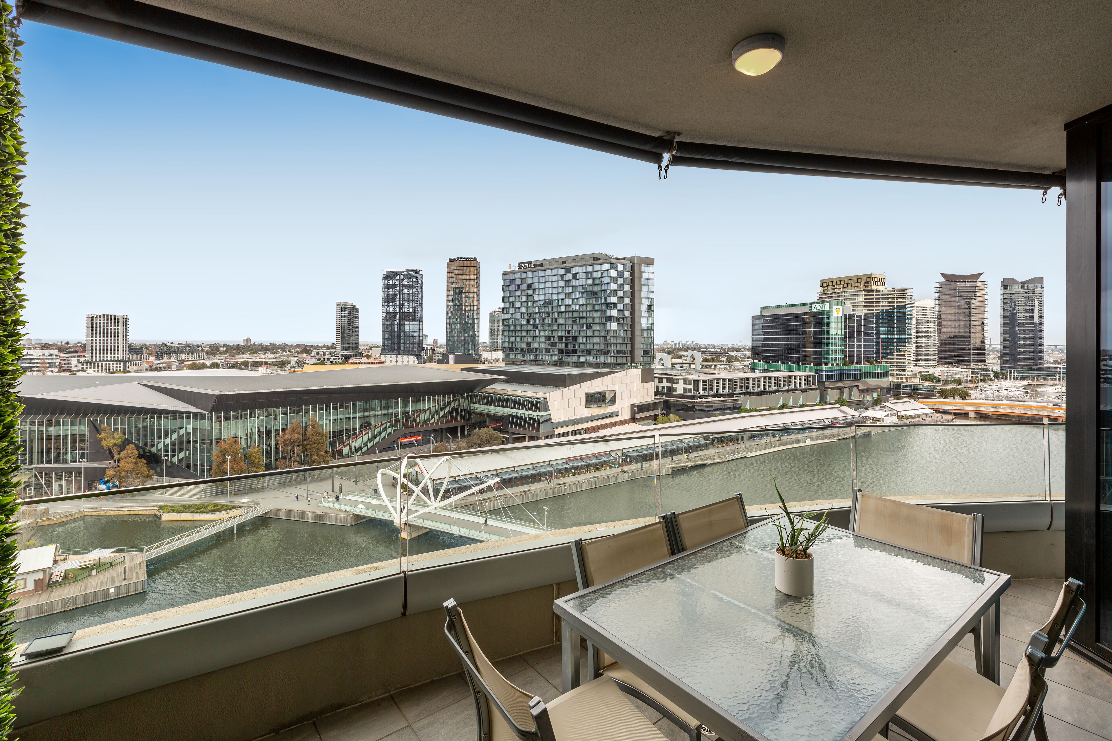A balcony with a glass table and chairs, overlooking a cityscape with modern buildings and a river.