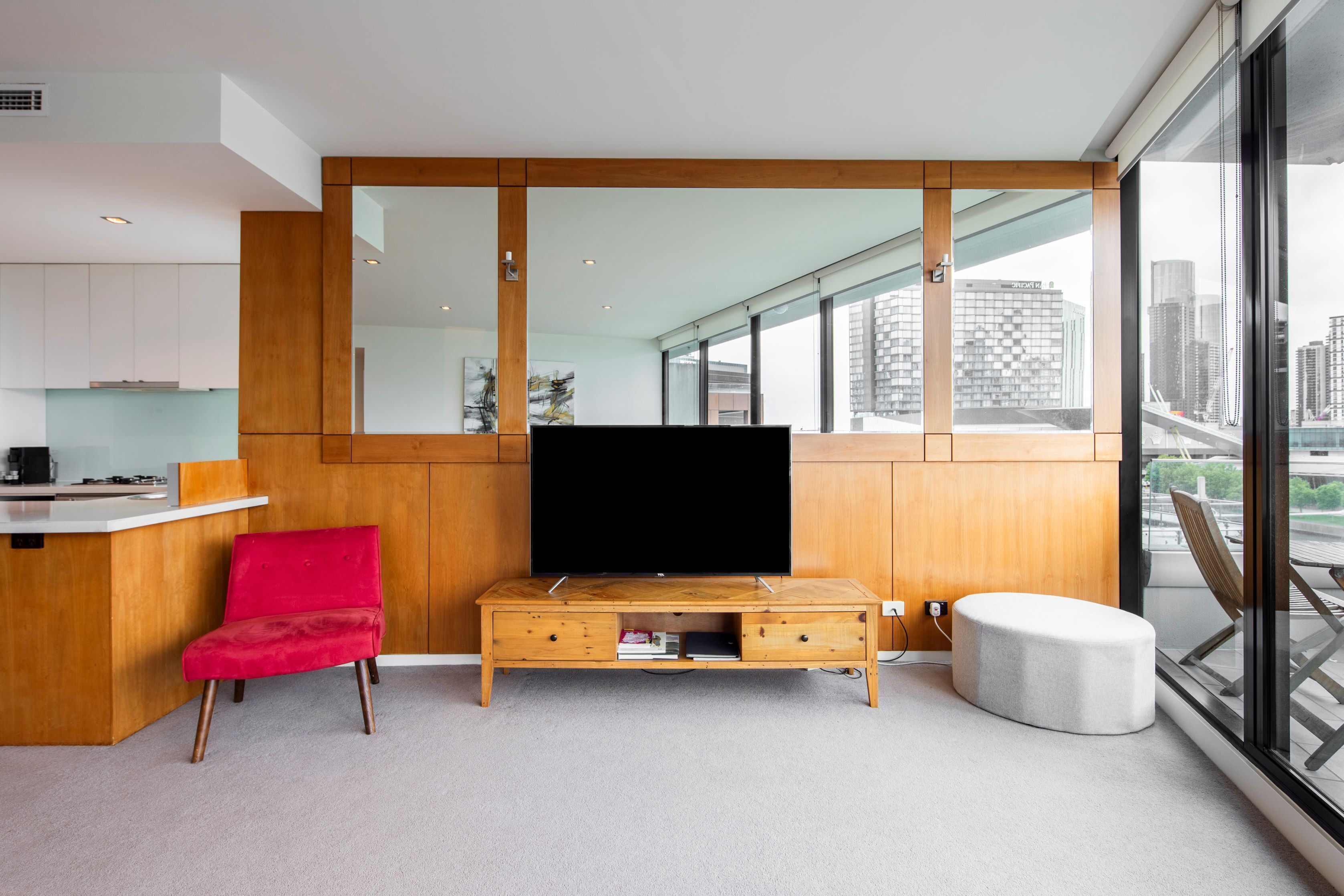 Un salon moderne doté d’un grand téléviseur à écran plat, de meubles en bois, d’un fauteuil rouge et offrant une vue sur la ville grâce à de grandes fenêtres.
