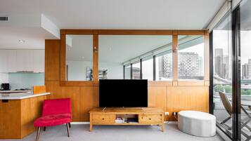 Un salon moderne doté d’un grand téléviseur à écran plat, de meubles en bois, d’un fauteuil rouge et offrant une vue sur la ville grâce à de grandes fenêtres.