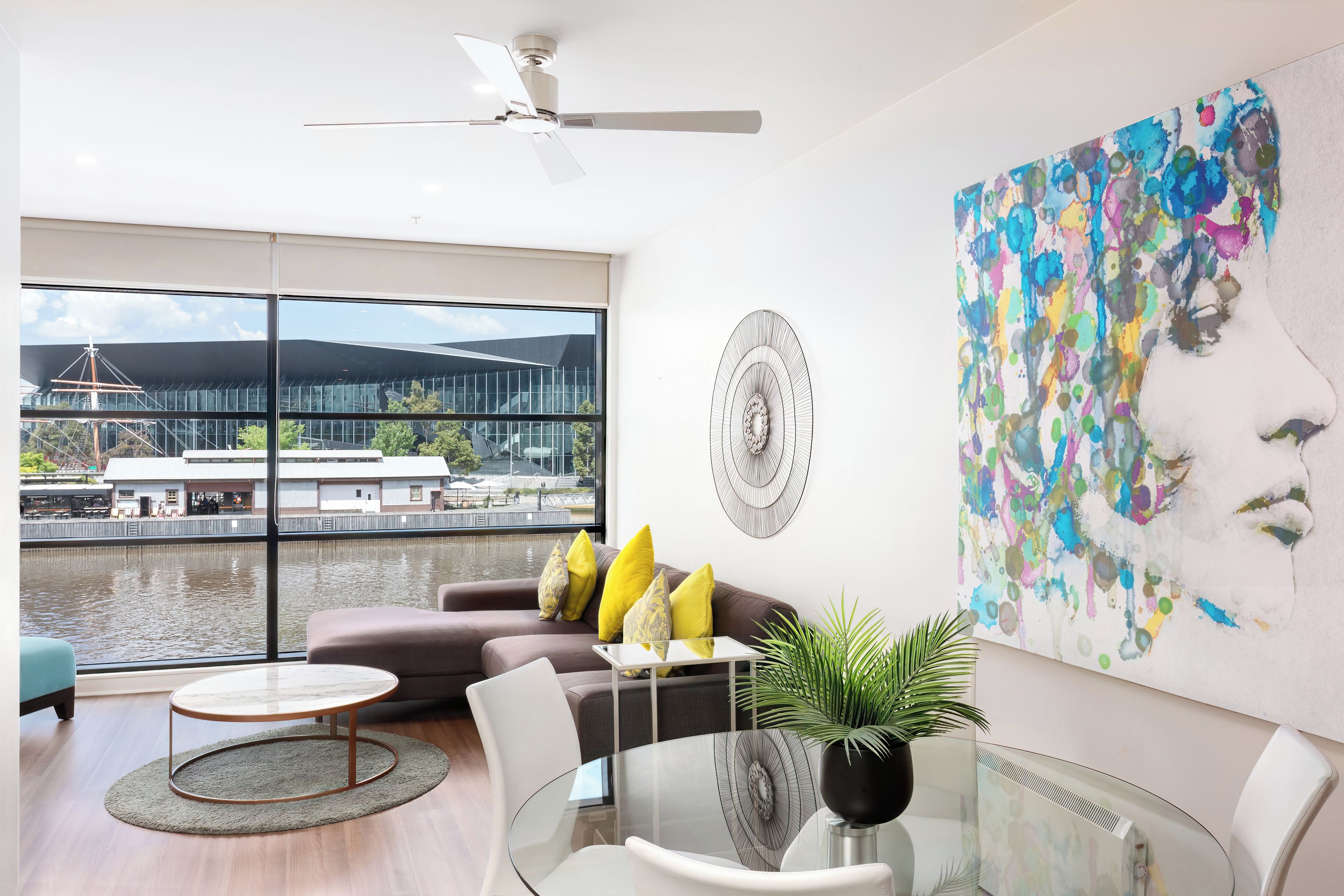 A modern living room with a view of a building across a body of water, a ceiling fan, and a large abstract painting.