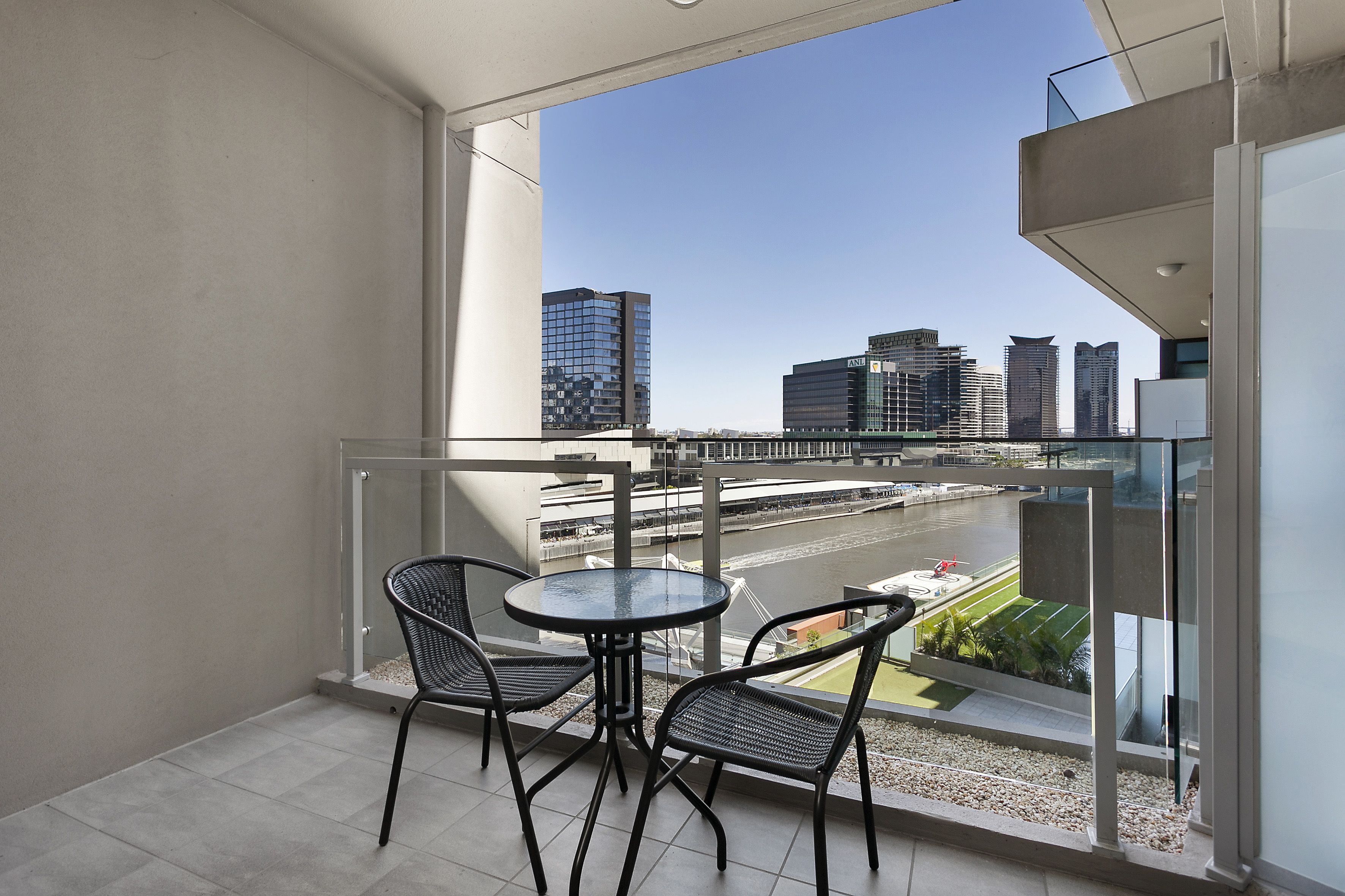 A balcony with a glass railing, two chairs, and a small table. A cityscape with buildings and a river is visible in the background.