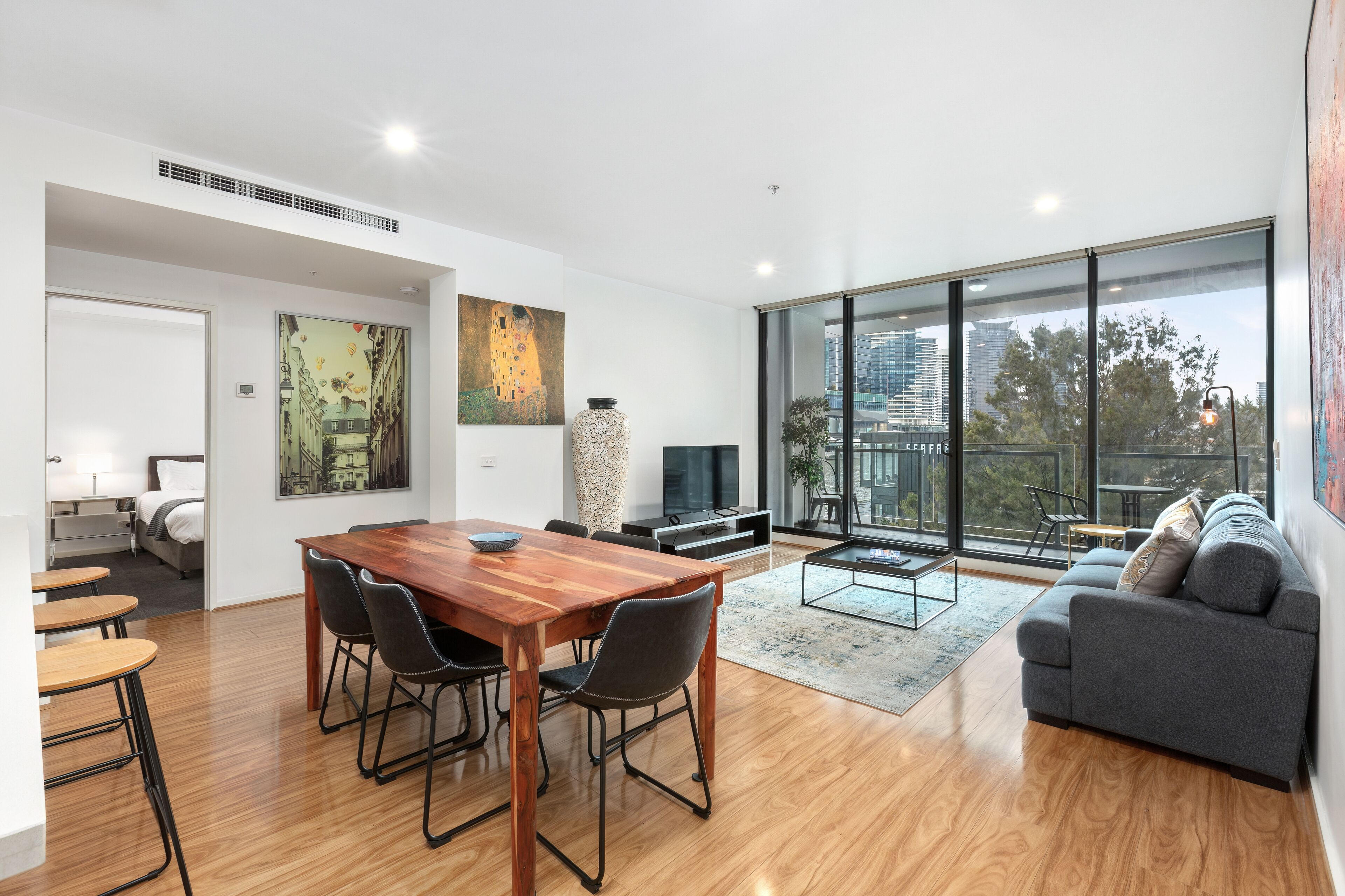 Una sala de estar moderna con una mesa de comedor de madera, un sofá gris y grandes ventanas que ofrecen vistas a la ciudad.