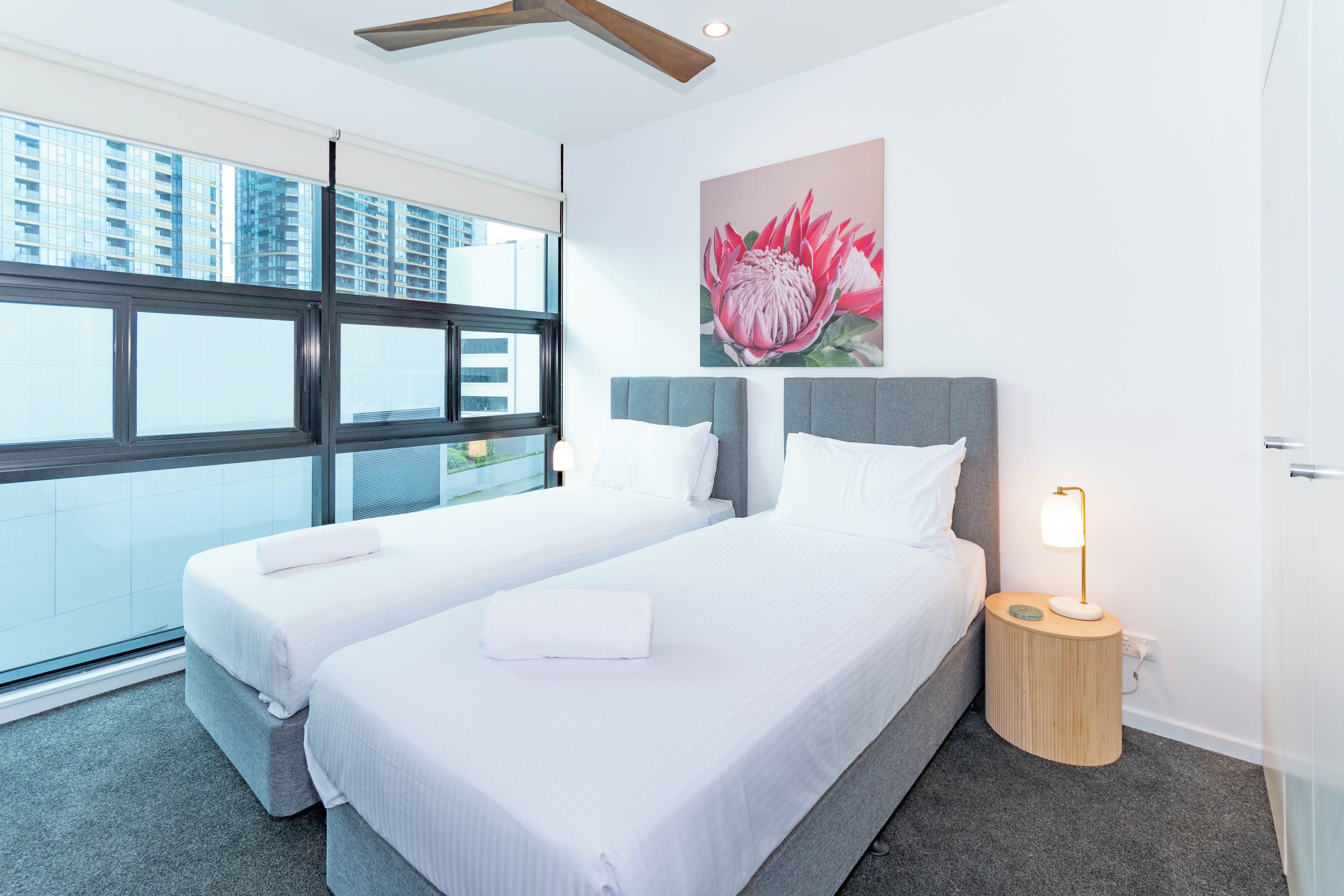 A hotel room with two beds, a large window, a painting of a flower, and a bedside lamp.