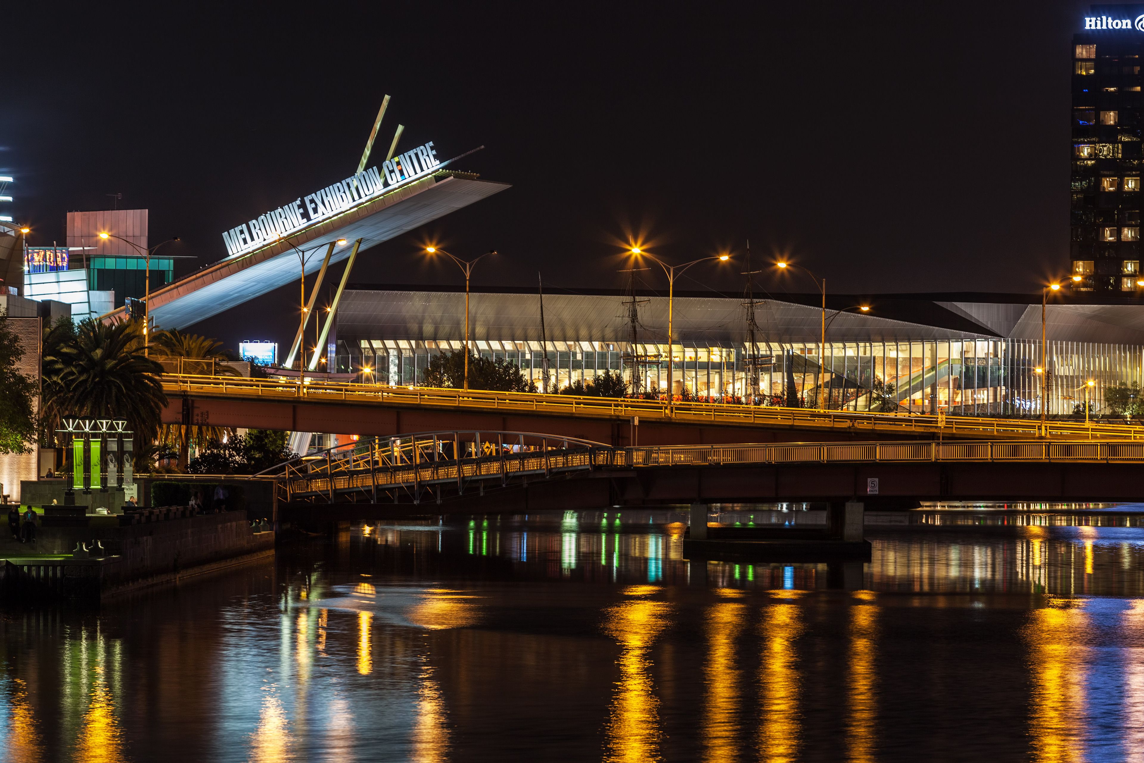 A modern bridge with a sign reading "Melbourne Exhibition Centre" and a Hilton hotel in the background.