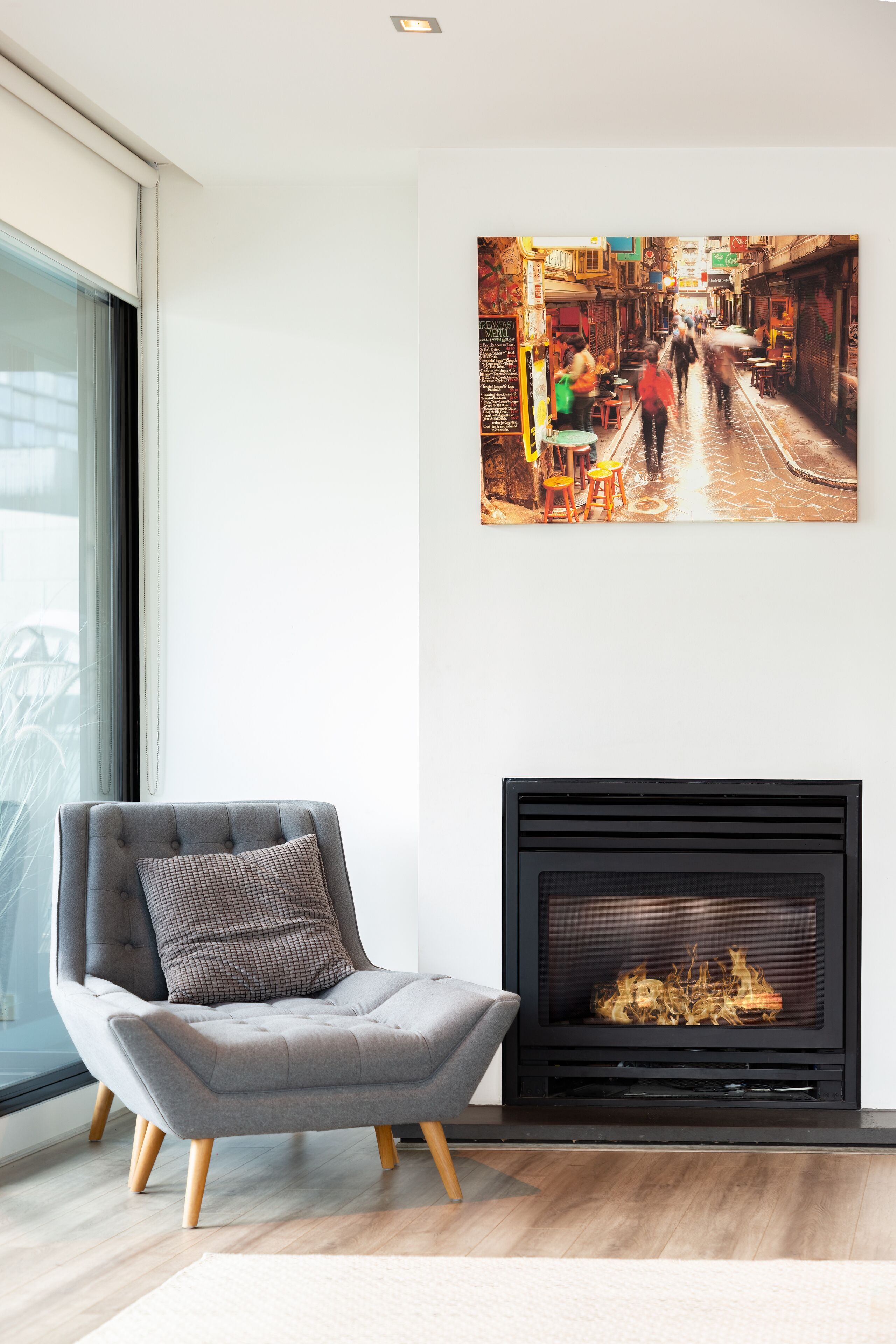 A modern living room with a fireplace, a comfortable armchair, and a large framed artwork of a bustling street scene.