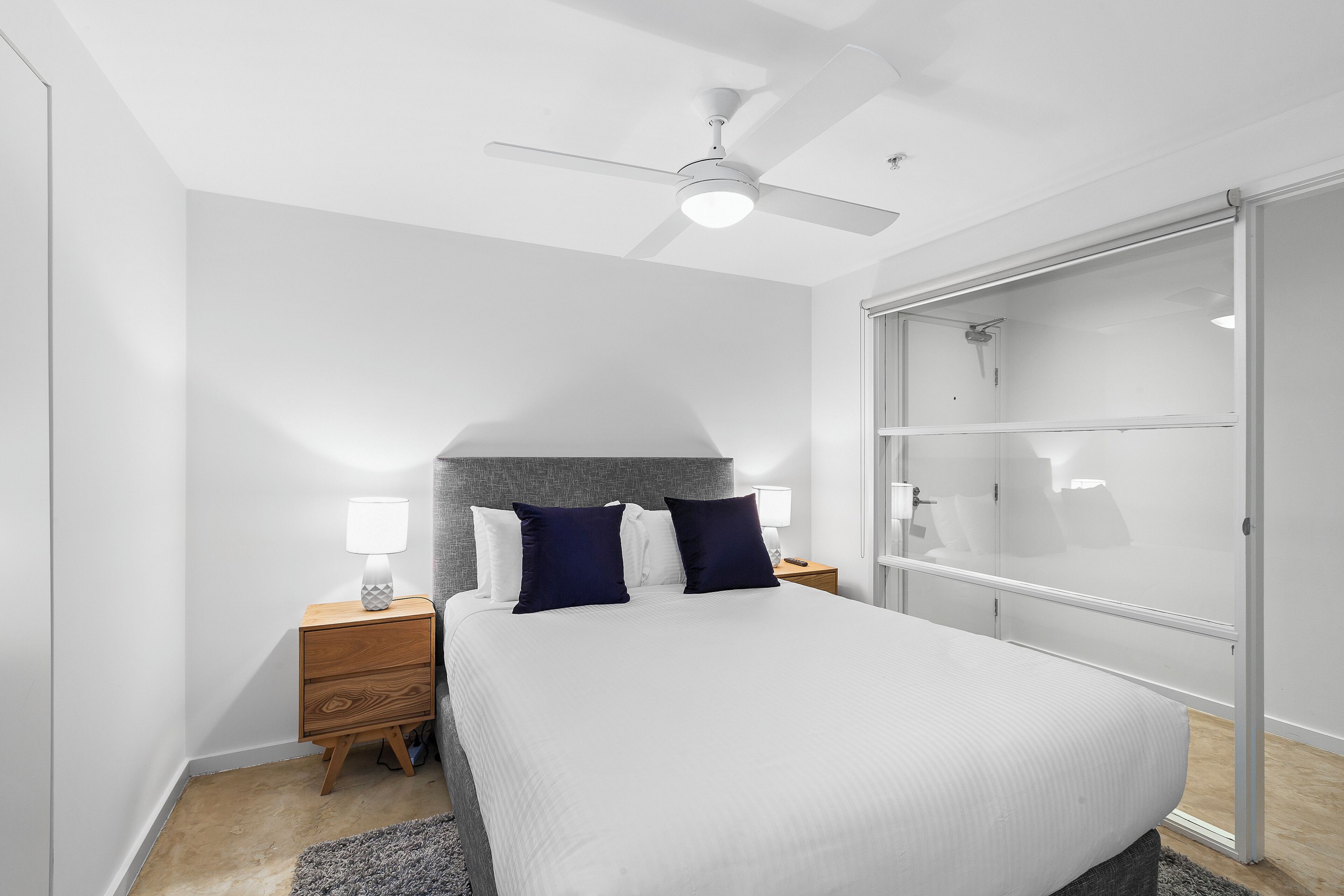Une chambre à coucher comprenant un lit, des tables de chevet, un ventilateur de plafond et un placard avec une porte coulissante en verre.