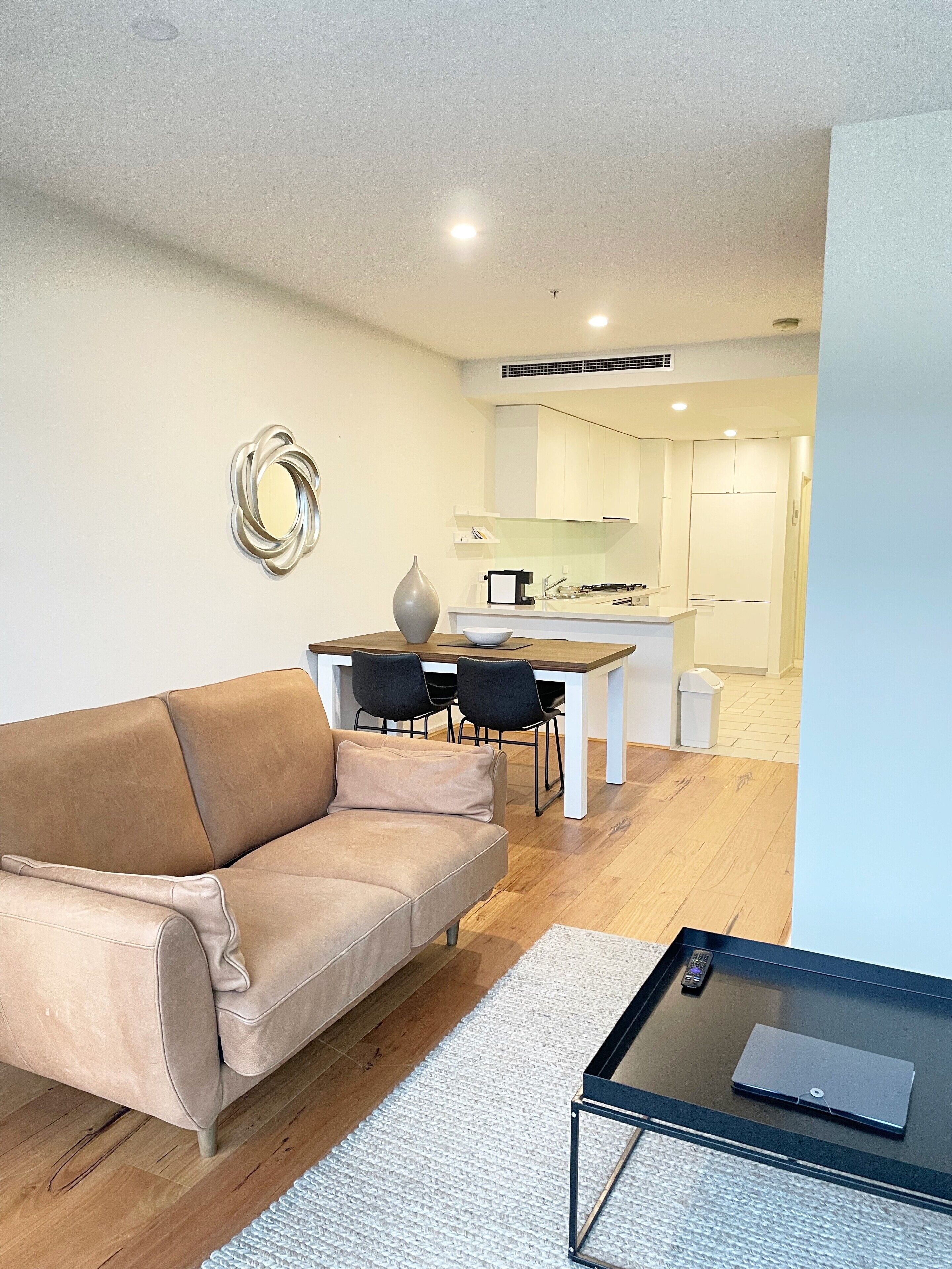Una sala de estar moderna con un sofá color beige, una mesa de comedor con sillas, una pequeña cocina y una mesa de centro de vidrio.