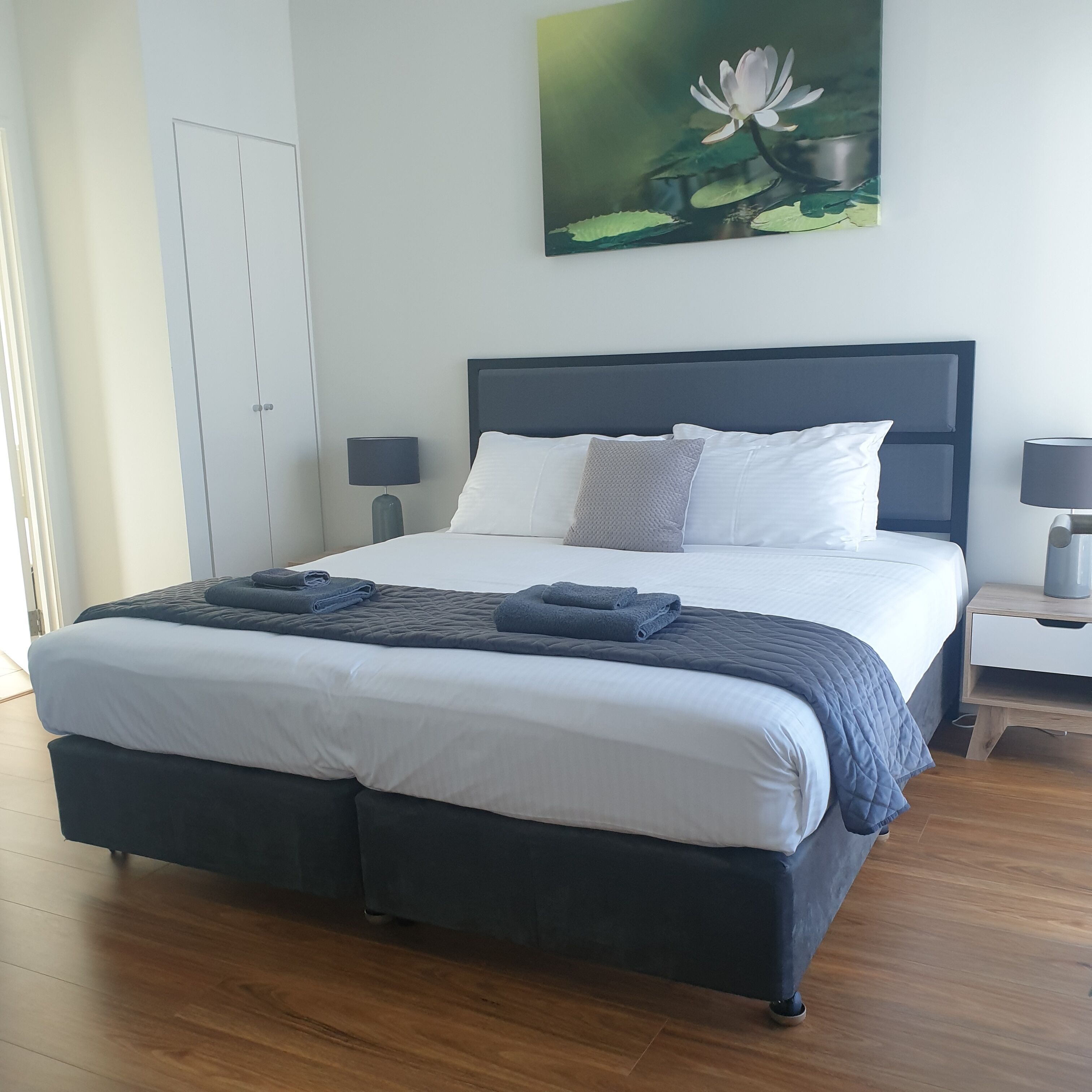 A neatly made bed with a dark headboard, white bedding, and two gray pillows. There are two bedside tables with lamps, a wooden floor, and a painting of a lotus flower on the wall.